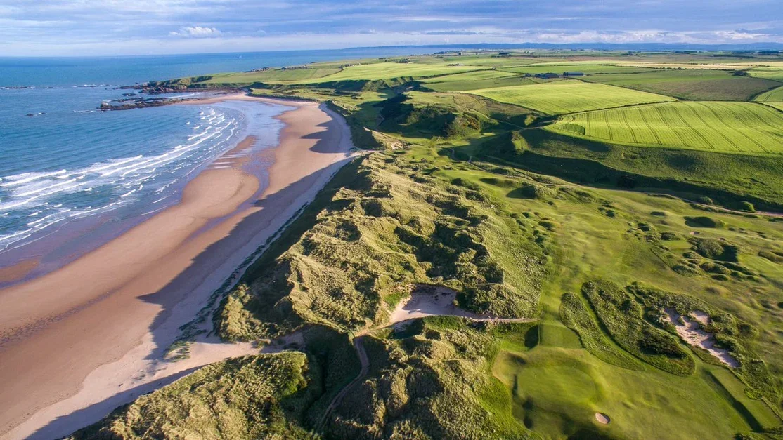 Aerial view of a coastline with a sandy beach, green hills, and farmland extending to the horizon under a partly cloudy sky.