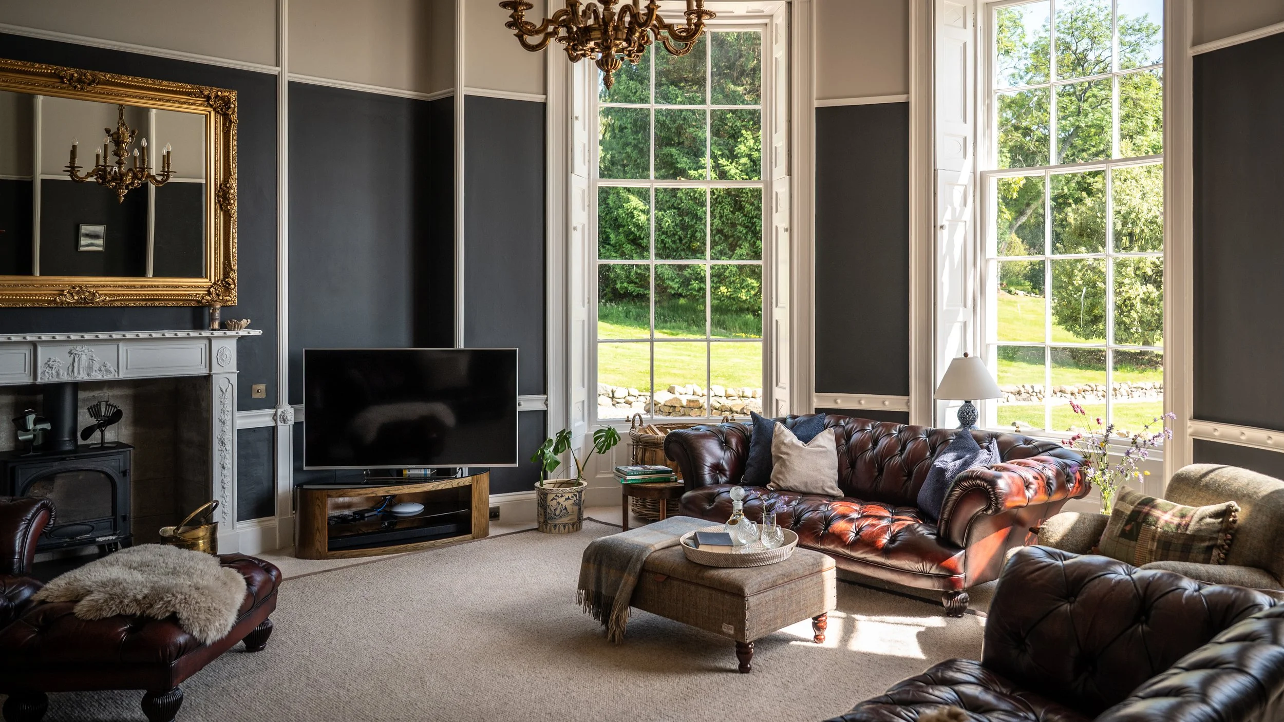 Living room with large windows showing greenery outside, dark walls with white trim, a fireplace with decorative mantle, a mounted flat-screen TV, a leather sofa with pillows, and a chandelier hanging from the ceiling.