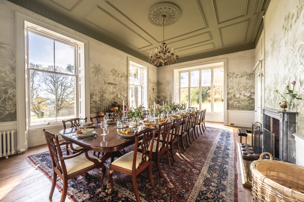 A formal dining room with a long wooden table set for a meal, surrounded by wooden chairs with upholstered seats, large windows allowing natural light, floral wallpaper and a decorative chandelier hanging from the ceiling.