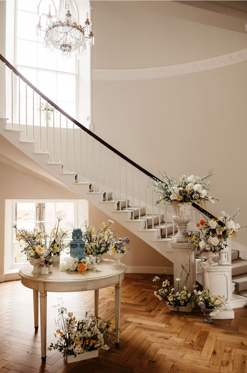Elegant interior with a staircase decorated with white and colorful flower arrangements, a round table with floral displays and a blue cake, and a chandelier hanging from a large window.