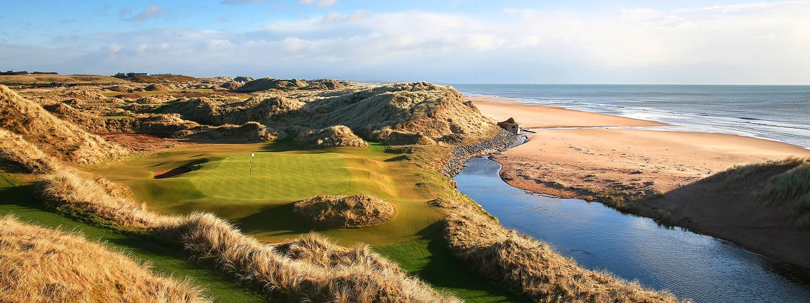 A coastal golf course with green fairways and sand dunes next to a sandy beach and ocean under a partly cloudy sky.