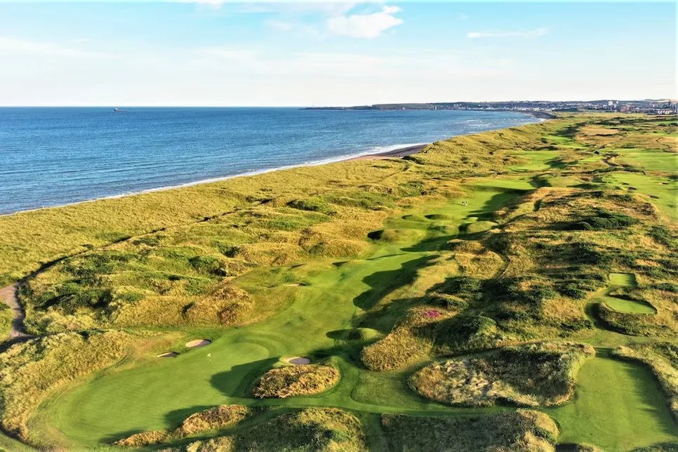 Aerial view of a golf course with rolling greens and sand traps along a coastline with the ocean in the background.