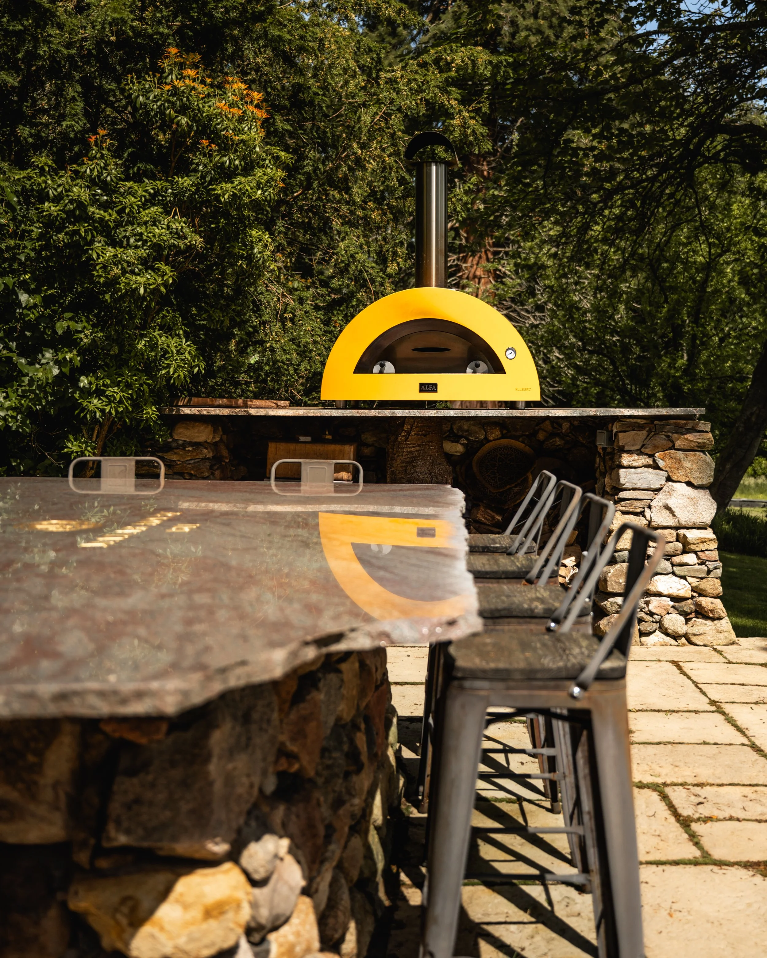 Outdoor scene showing a row of chairs along a stone wall, with a pizza oven and a green, leafy tree in the background.