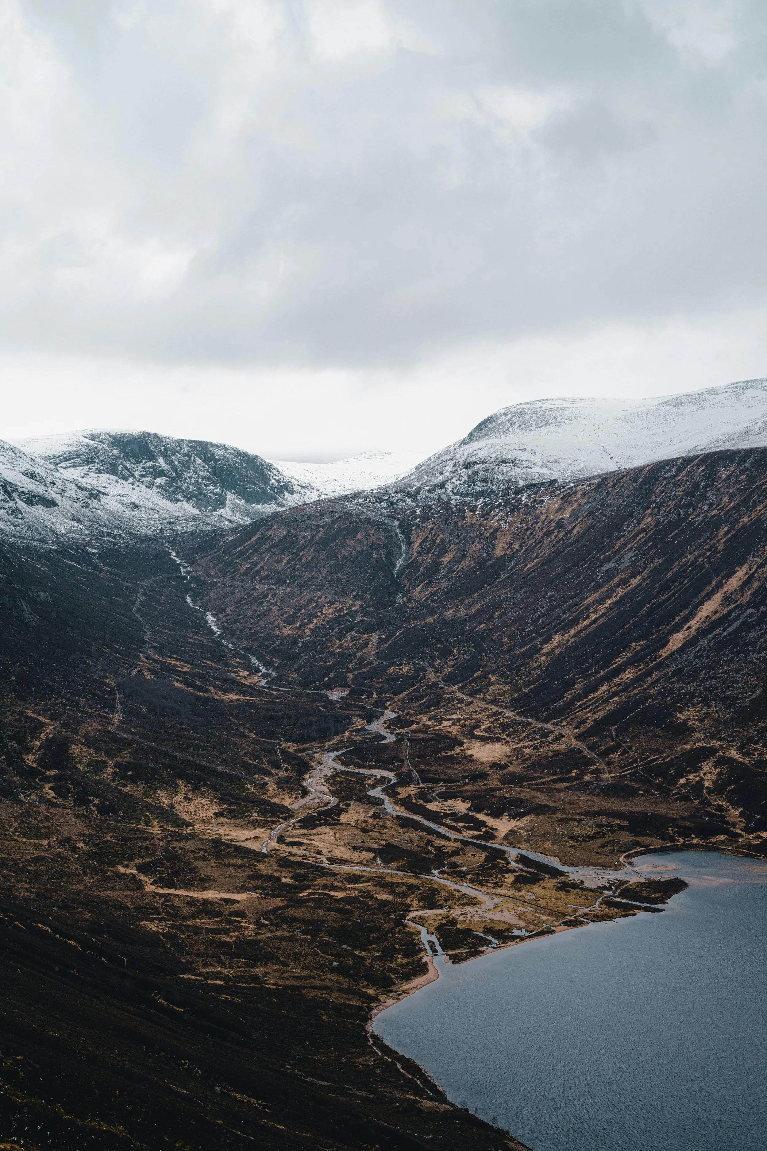 Snow-capped mountains overlooking a river valley with winding streams, a shoreline, and a cloudy sky.