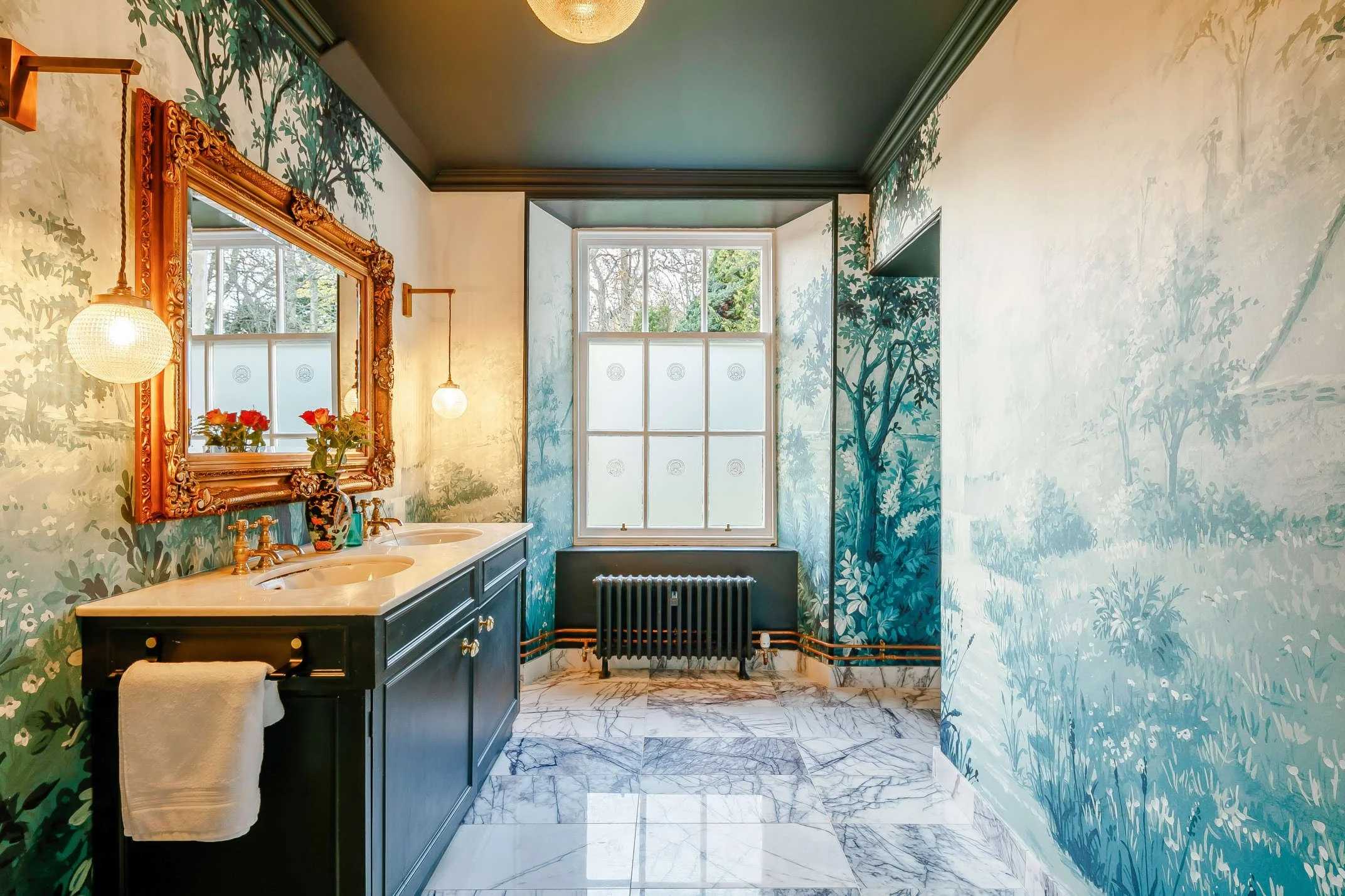 Bathroom with nature-inspired wallpaper murals, a double vanity with brass fixtures and a large ornate mirror, a window with frosted glass, a black radiator, and a marble floor.