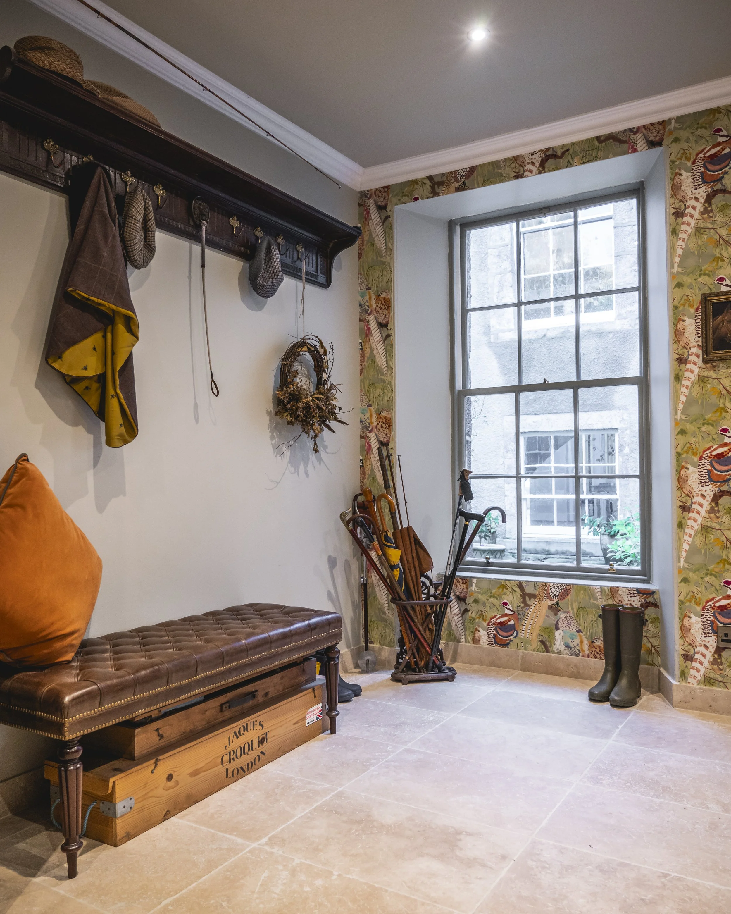 A cozy interior space with a large window, a cushioned bench, a wooden crate, and various decorative items on the wall including coats, hats, and a wreath.
