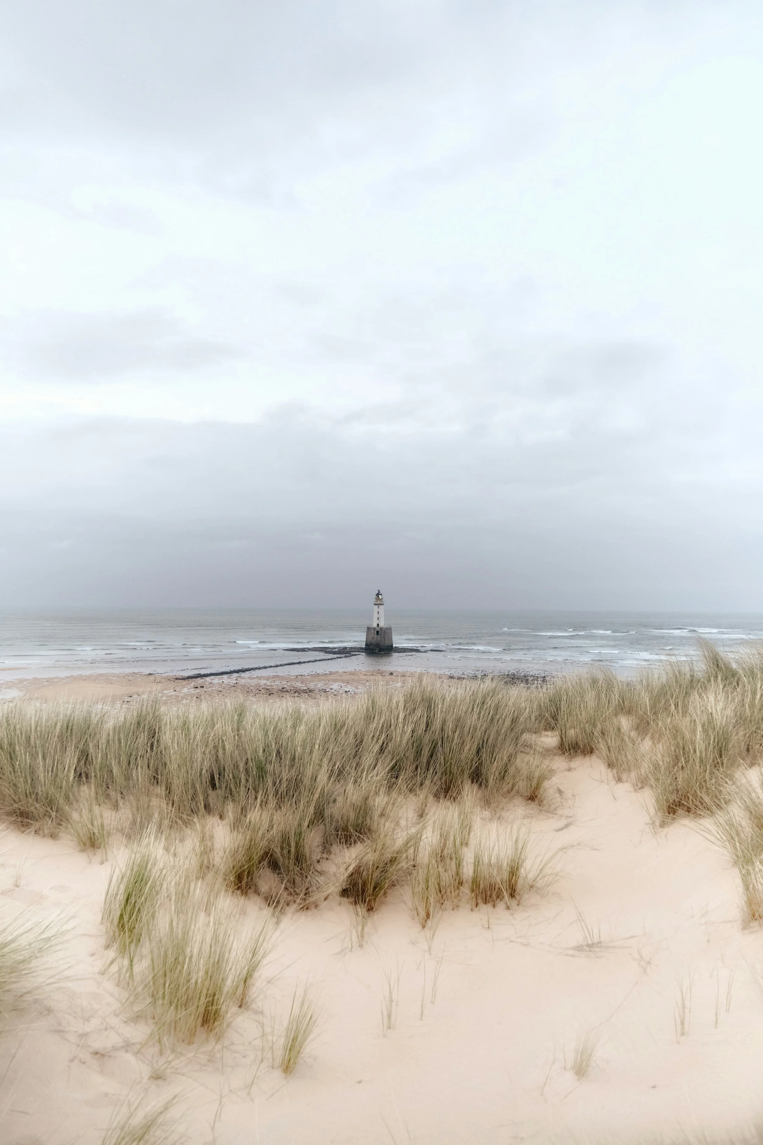 A lighthouse on a rocky shoreline seen from sandy beach with tall grass, cloudy sky over the ocean.