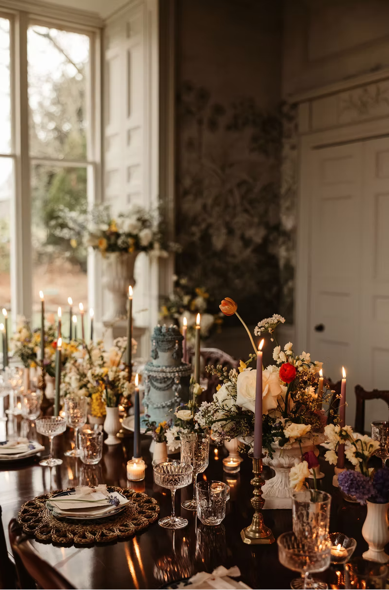 A beautifully decorated dining table with candles, floral arrangements, and fine china, set in a room with large window and floral wallpaper.