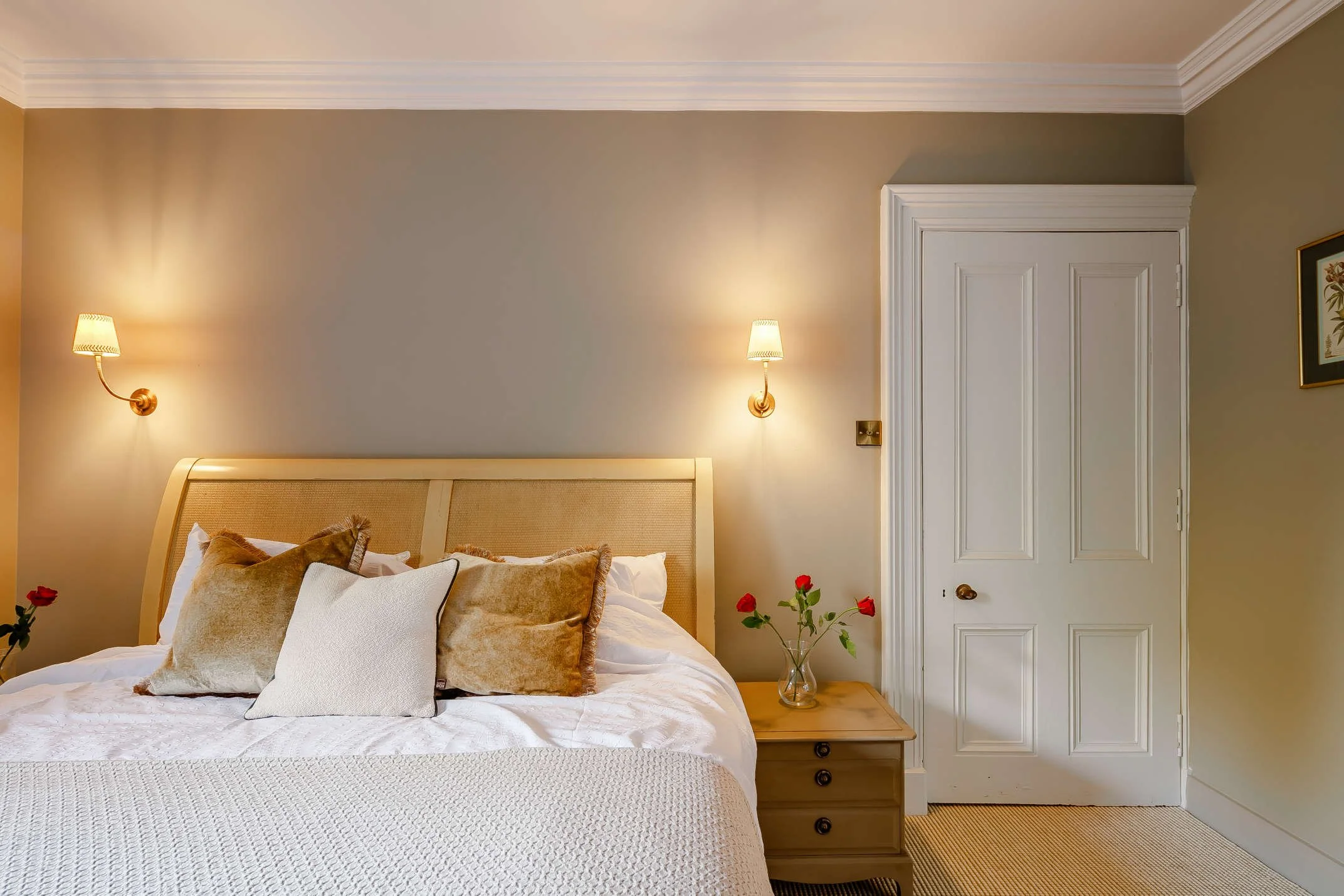 A cozy bedroom with a bed, multiple pillows, bedside table with a vase of red roses, yellow walls, white door, and wall-mounted lamps.
