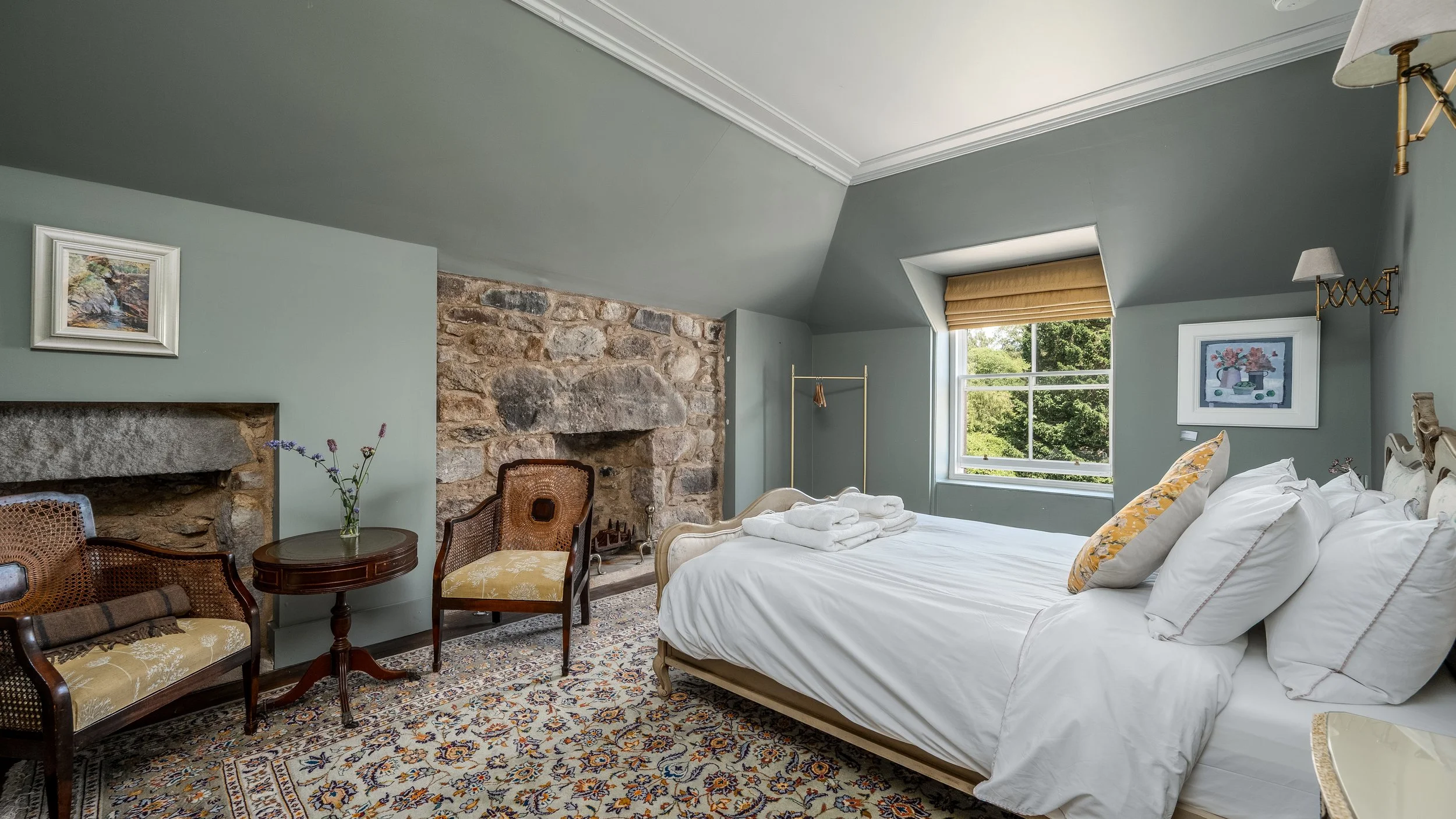 A cozy bedroom with a large window showing green trees outside. The room has a stone fireplace, a bed with white linens and yellow pillows, and vintage chairs around a small table with a vase of flowers. There are framed paintings on the walls and a patterned area rug on the floor.