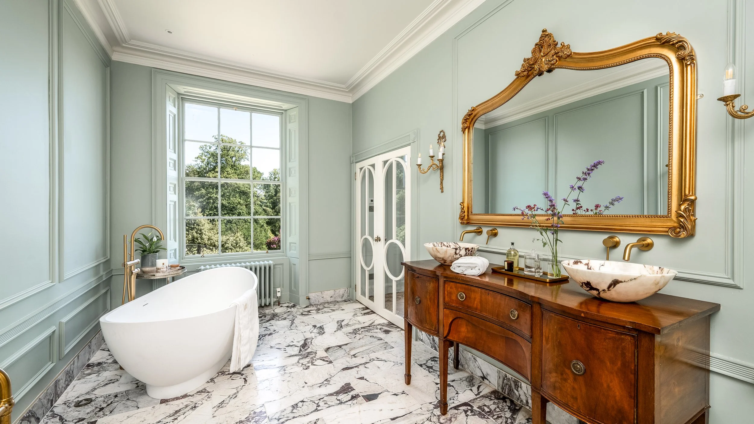 Elegant bathroom featuring a large window with a view of trees, a clawfoot bathtub with a towel, a vintage wooden vanity with two marble vessel sinks, a large ornate gold mirror, a gold wall sconce, and marble flooring.