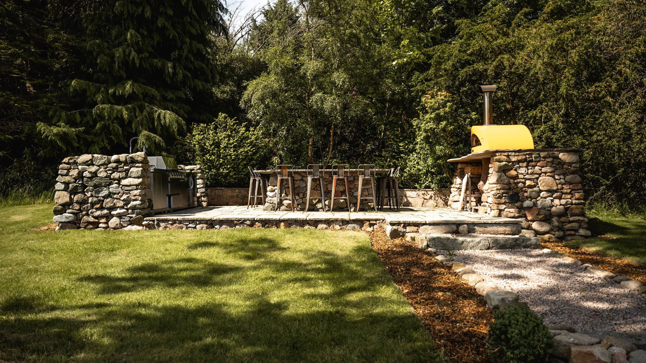 Outdoor stone patio with a stone and wood table, chairs, a yellow pizza oven, and a grill set in a grassy yard surrounded by trees.