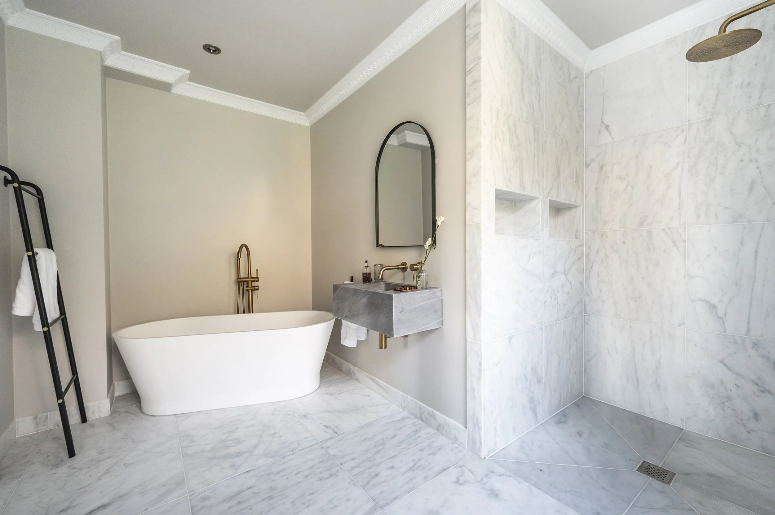 Modern bathroom with white freestanding bathtub, marble flooring, and a marble shower wall with niches, brass fixtures, a mirror, and minimalist decor.