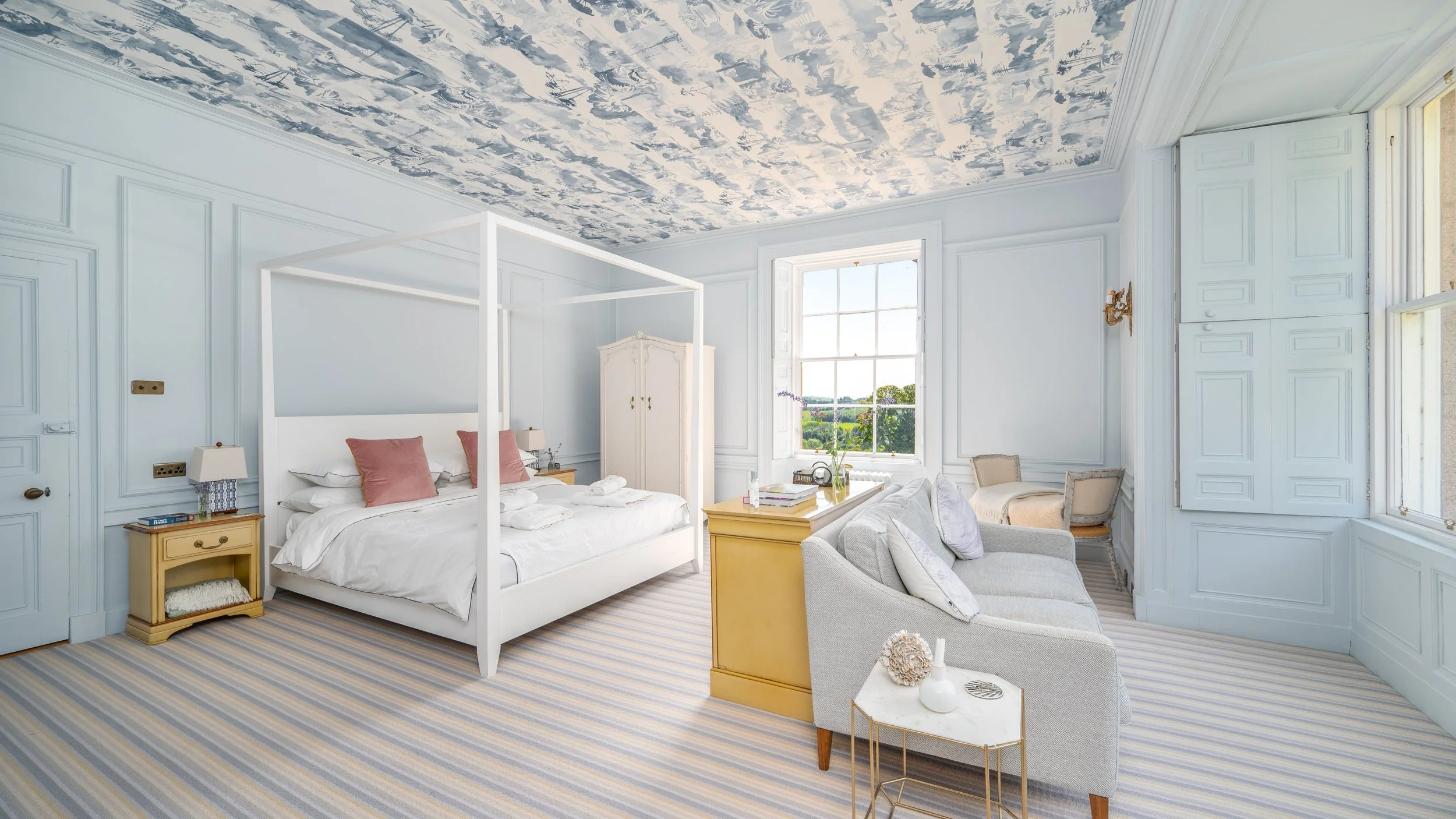 A bright bedroom with large windows showing a green landscape outside, a white bed with pink pillows, light-colored side tables, a cozy seating area, and a decorative ceiling pattern.