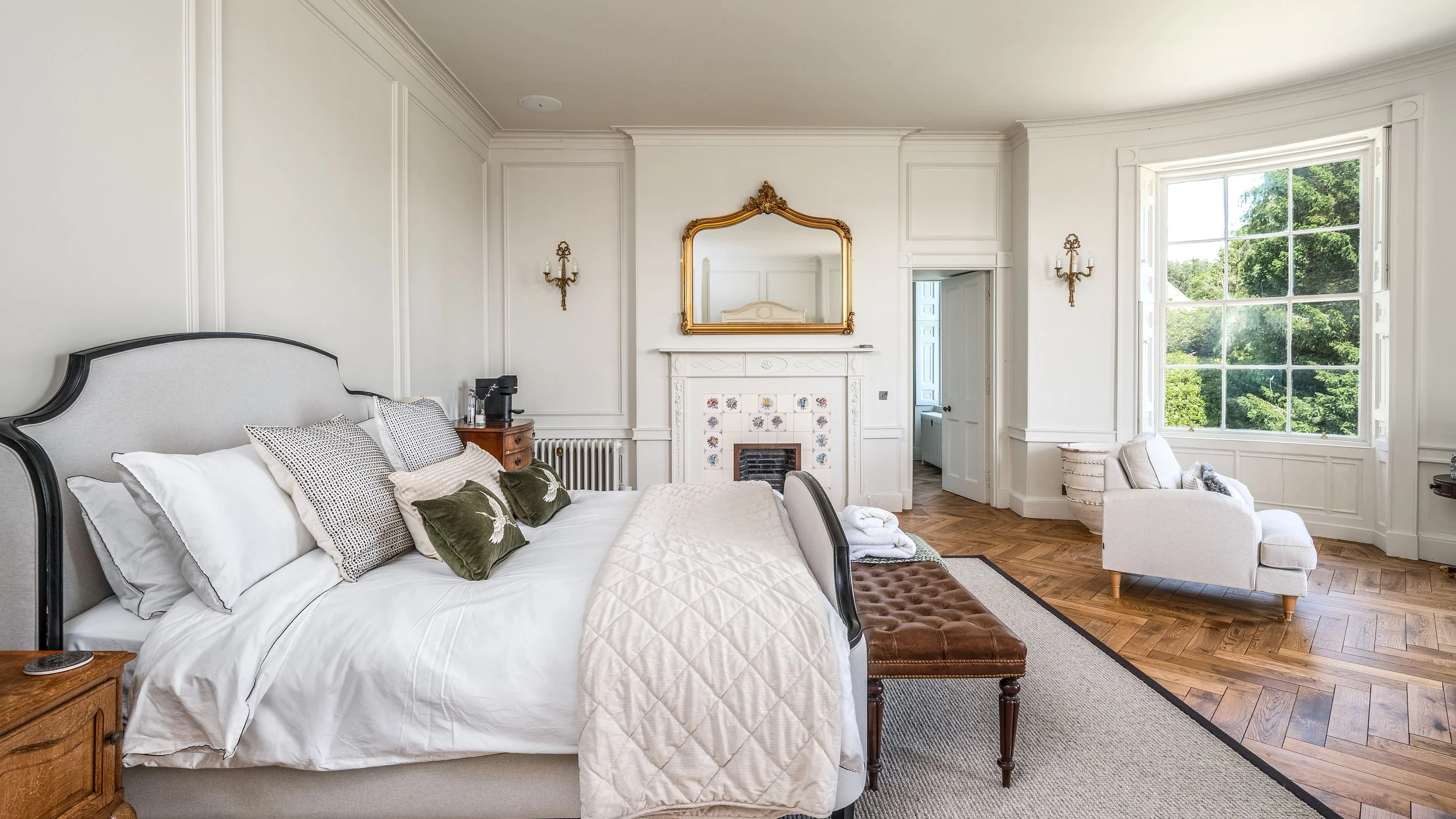 A bright bedroom with white walls, a large window with a view of green trees, a white bed with multiple pillows, a brown leather bench, a small armchair near the window, and a decorated fireplace with a gold-framed mirror above it.