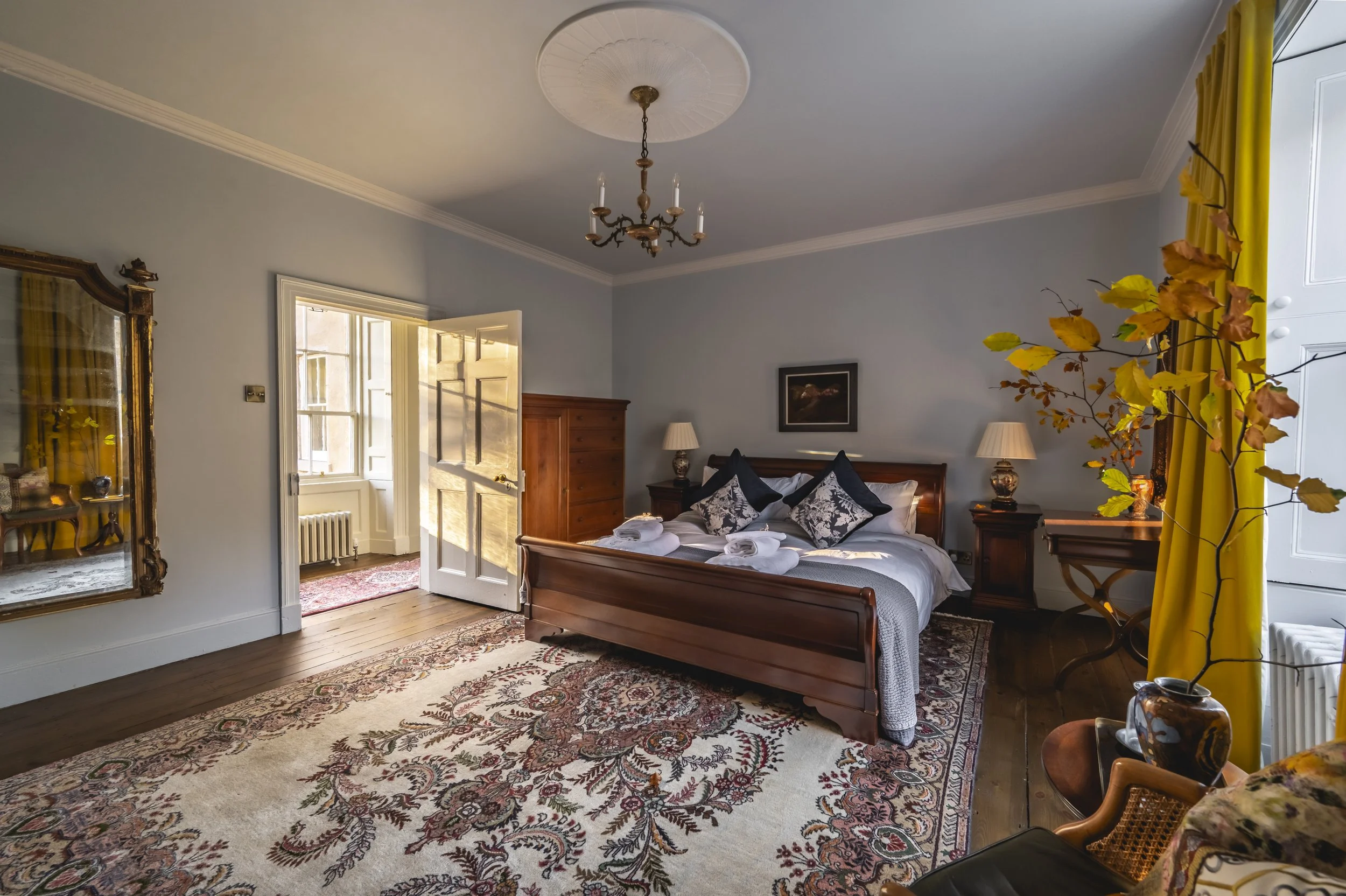 A bedroom with a wooden bed covered in white and dark blue pillows, two bedside lamps, a wooden dresser, and a picture on the wall. A large patterned rug covers the wooden floor, and yellow curtains frame a window.