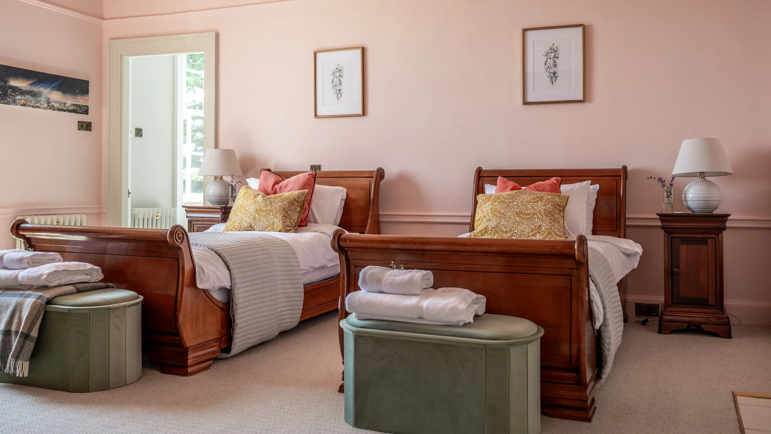 A cozy bedroom with two twin beds made of polished wood, each with pillows and bed coverings. Nightstands with lamps and vases with lavender are between the beds. Framed artwork hangs on the light pink walls, and a door opens to a balcony with greenery outside.
