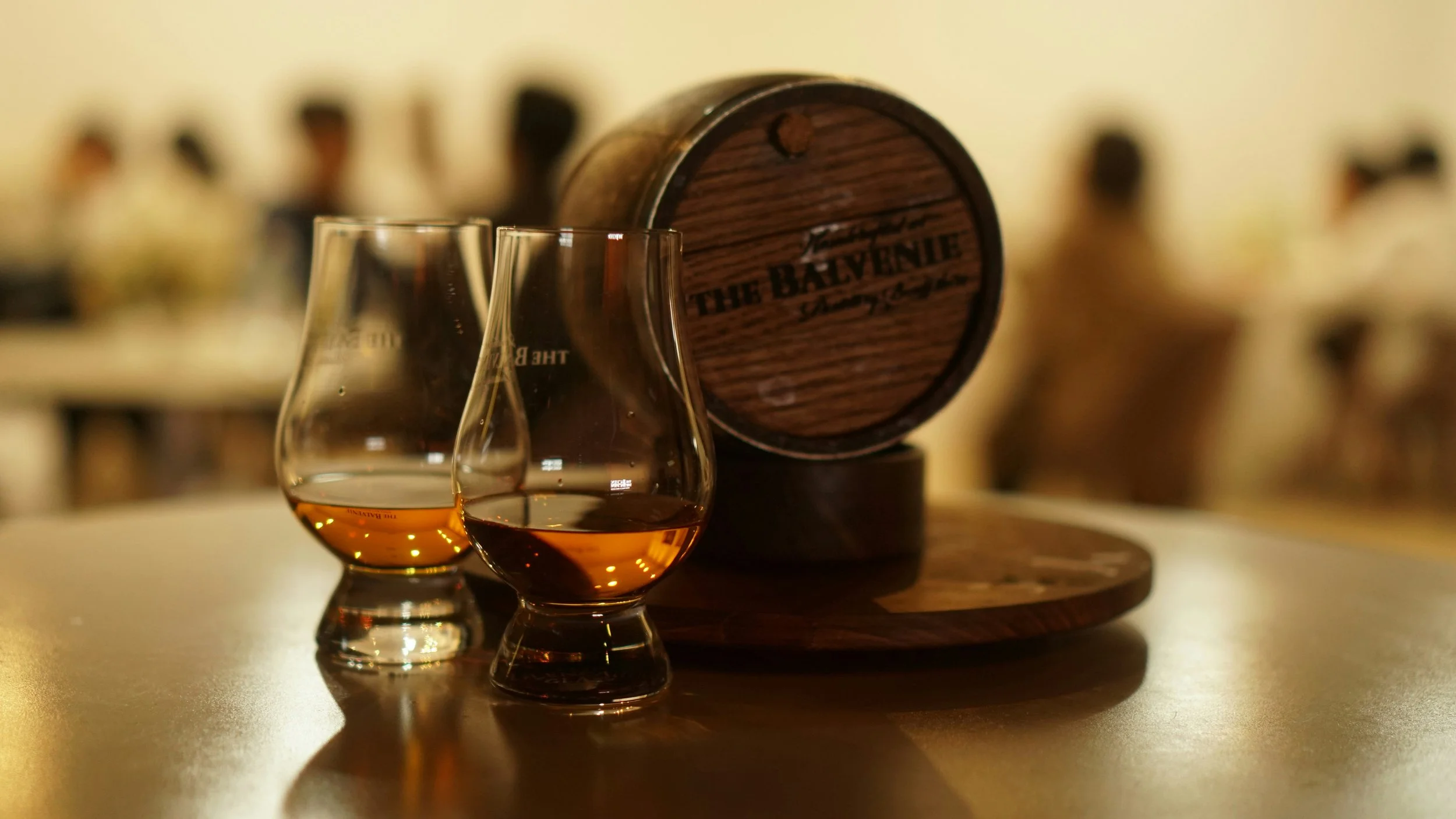 Two glasses of whisky or bourbon in front of a wooden barrel labeled 'The Balvenie'.