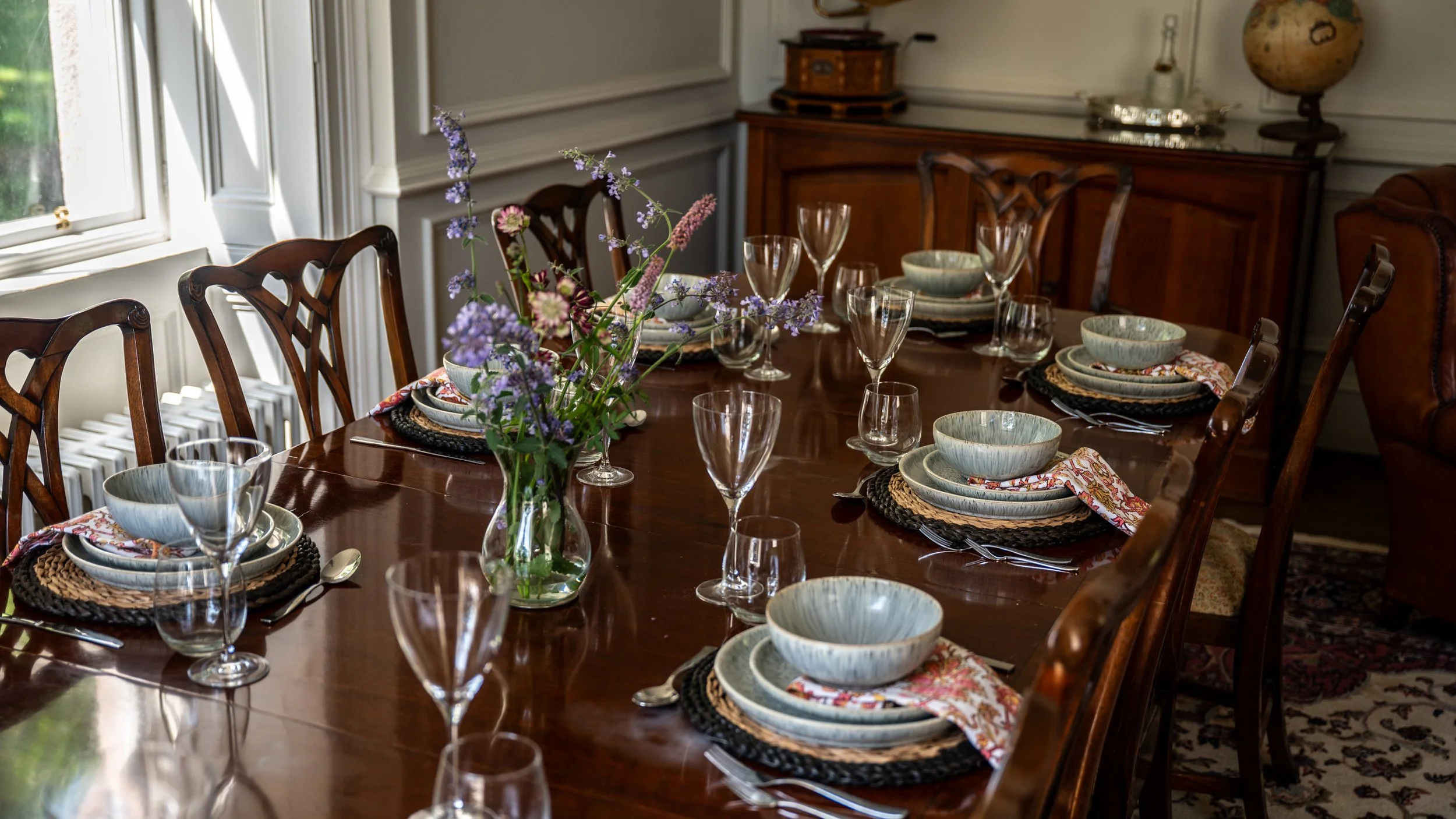 A formal dining room with a polished wooden table set with bowls, plates, silverware, and glasses. There is a centerpiece of purple wildflowers in a glass vase. The room has large windows, wooden chairs, and a sideboard with decorative items like a g