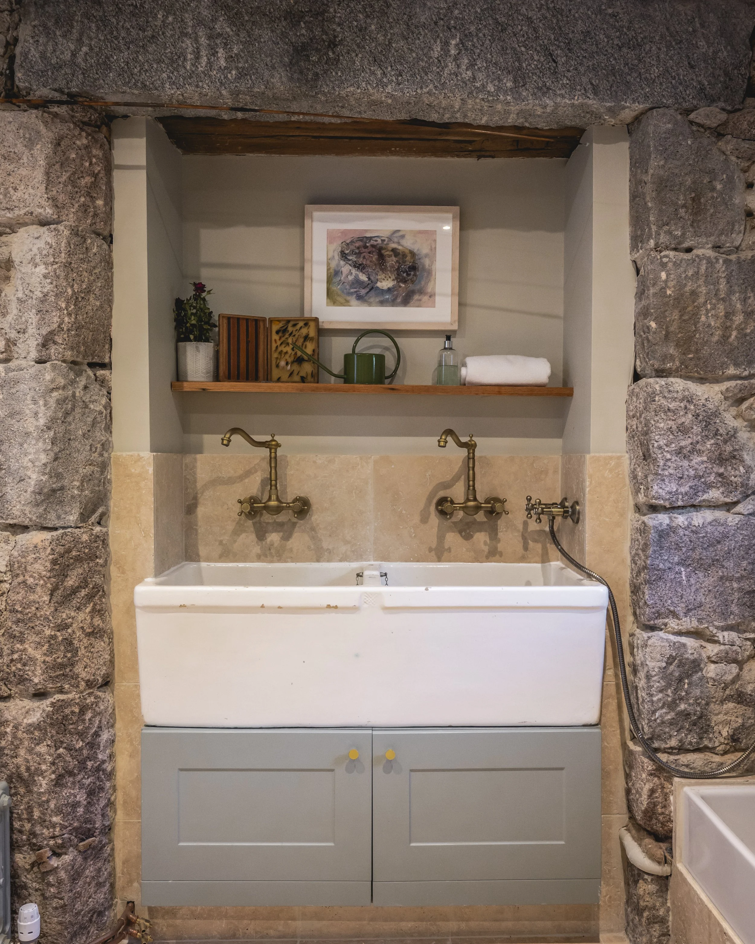 A rustic bathroom wash station with a white washing basin, brass vintage style faucets, a gray cabinet with yellow knobs, a wooden shelf above with a framed abstract artwork, a potted plant, watering can, and hand sanitizer, surrounded by stone walls