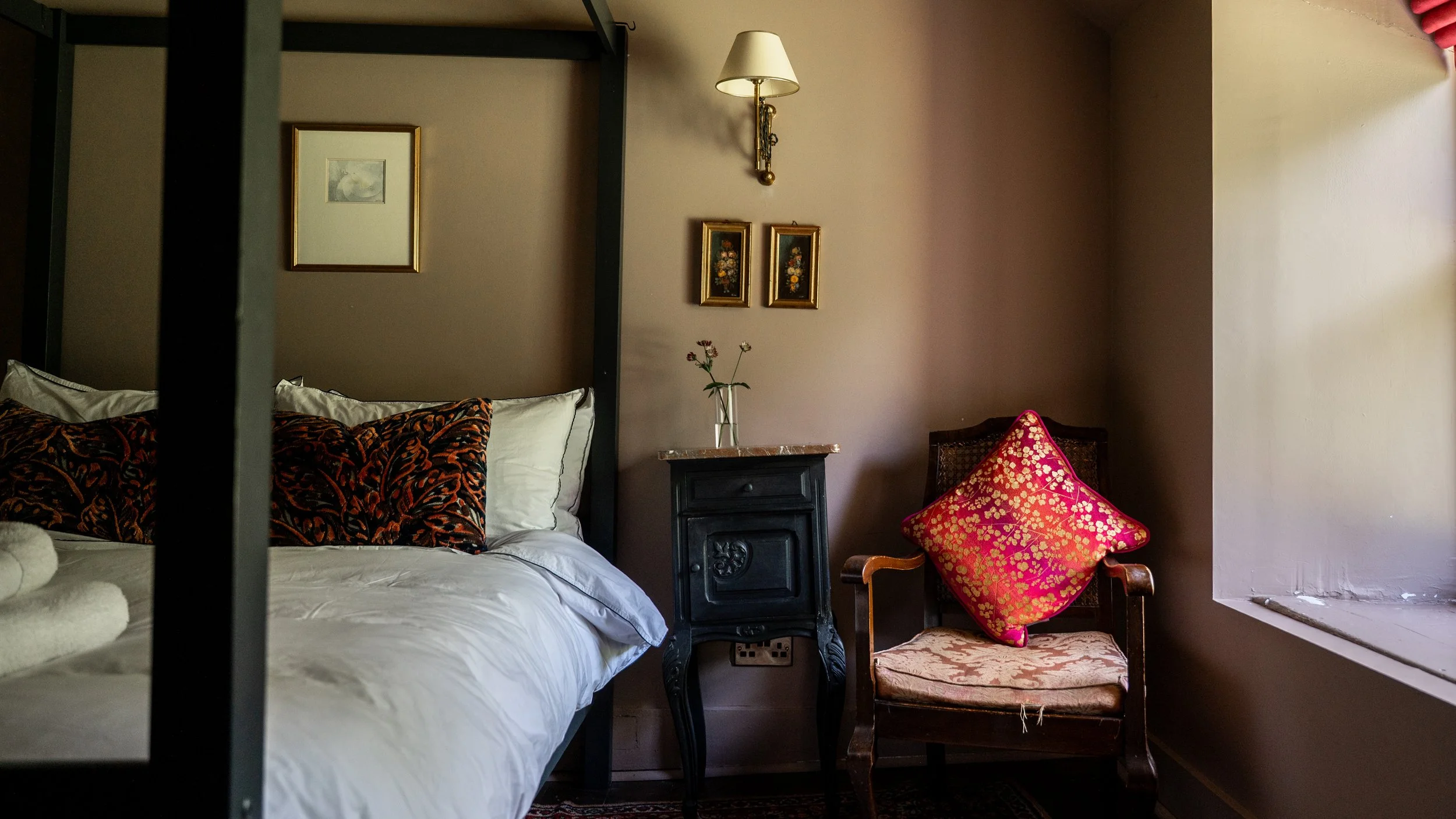 A cozy bedroom with a black metal bed frame, white bedding, a black nightstand with a glass vase and flowers, framed artwork on the wall, a wall-mounted lamp, a wooden chair with a pink and gold decorative pillow, and a window with red curtains.