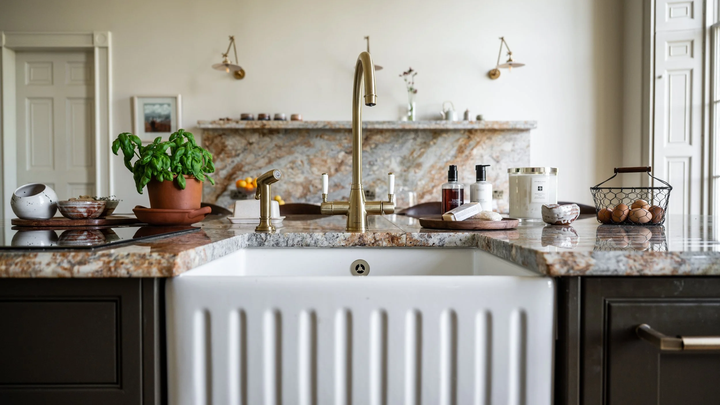 A kitchen with a granite countertop island, a gold faucet, and various items including a potted basil, bowls, soap dispensers, a jar, eggs in a wire basket, and a small soap dish.