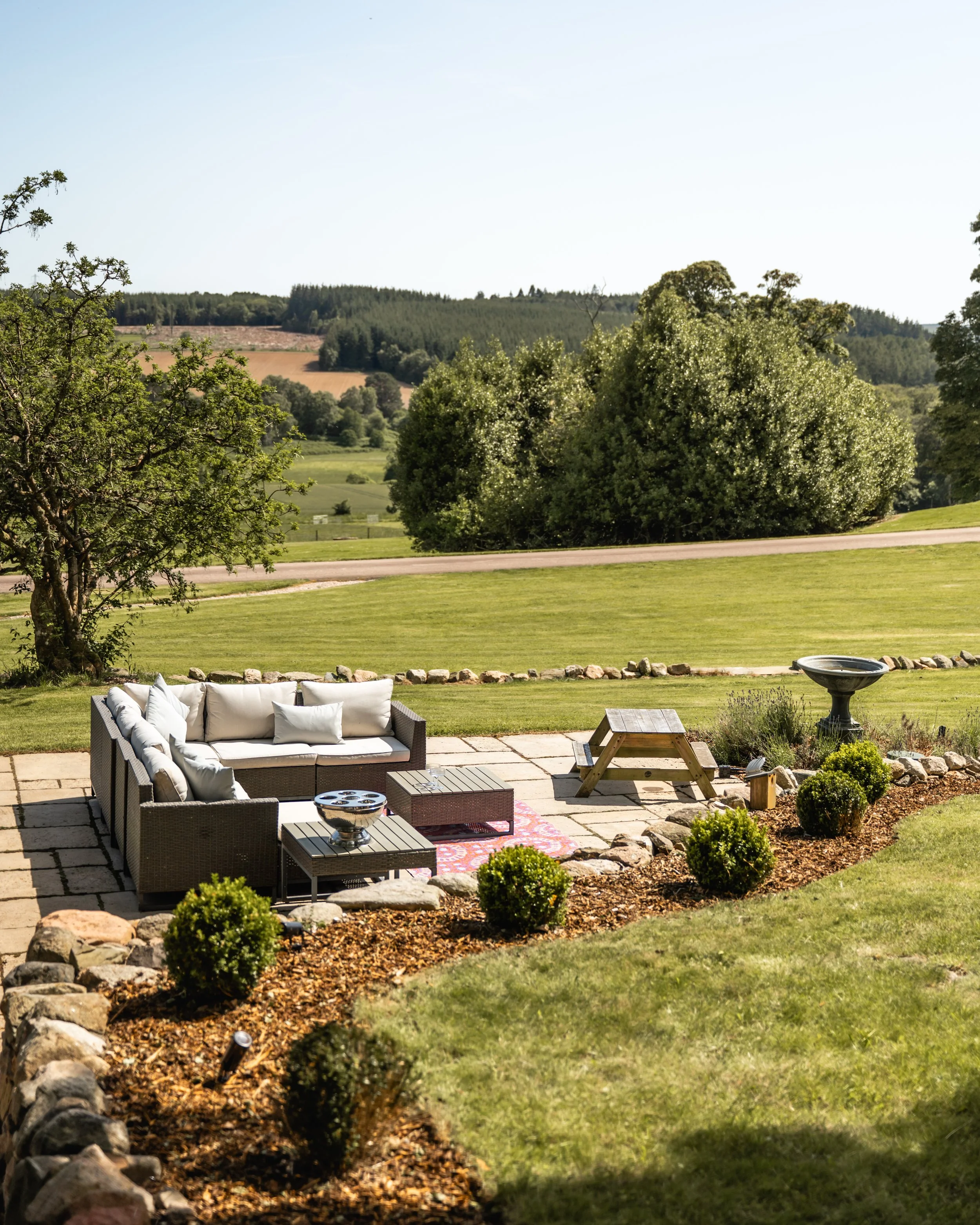 A backyard patio with outdoor wicker furniture, including a sectional sofa with white cushions, a side table, surrounded by a garden bed with small shrubs and a birdbath, overlooking rolling hills and farmland.