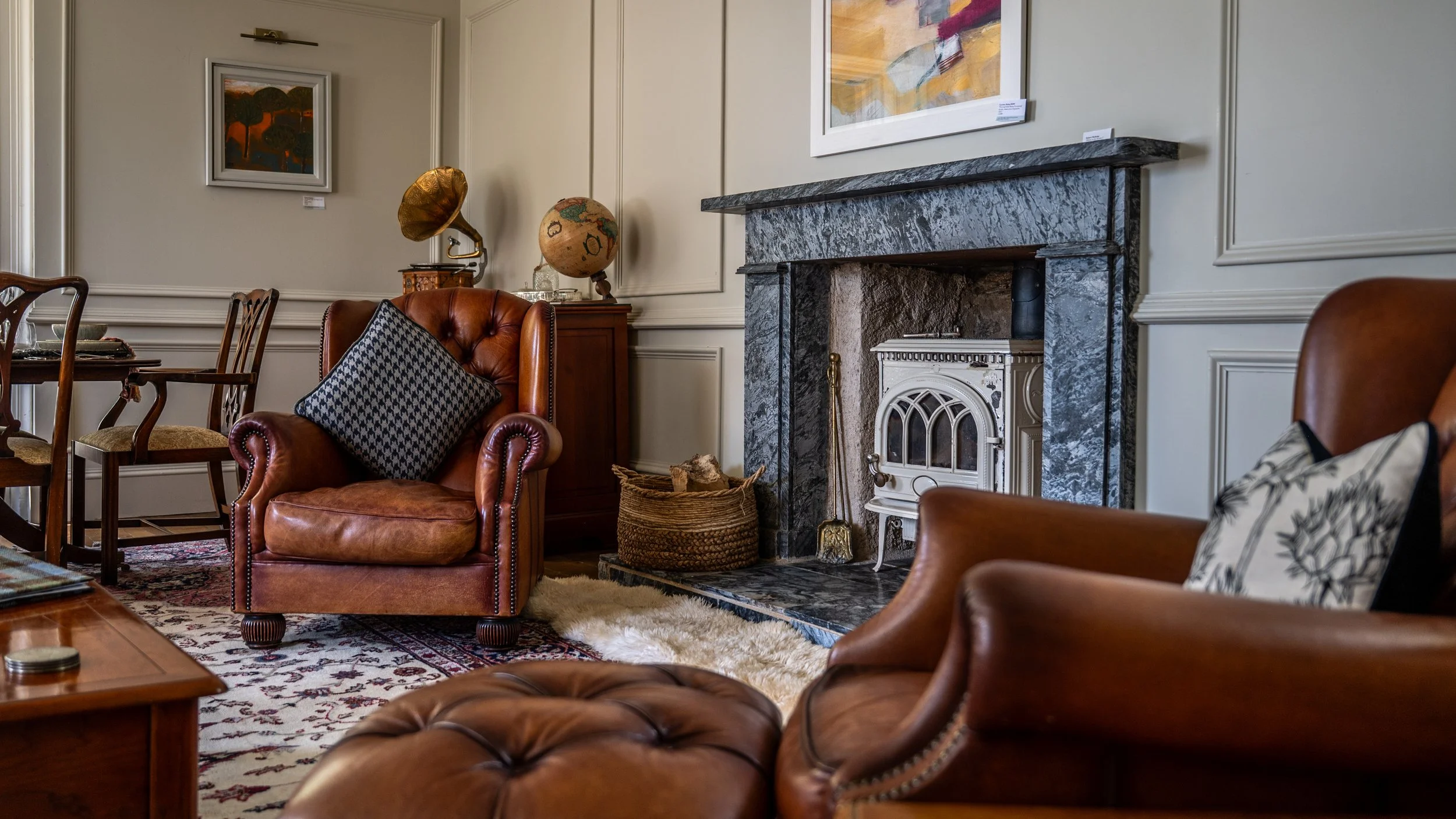 A cozy living room with a fireplace, leather armchairs, wooden dining table with chairs, abstract paintings on the wall, and decorative items like a vintage globe and a gramophone.