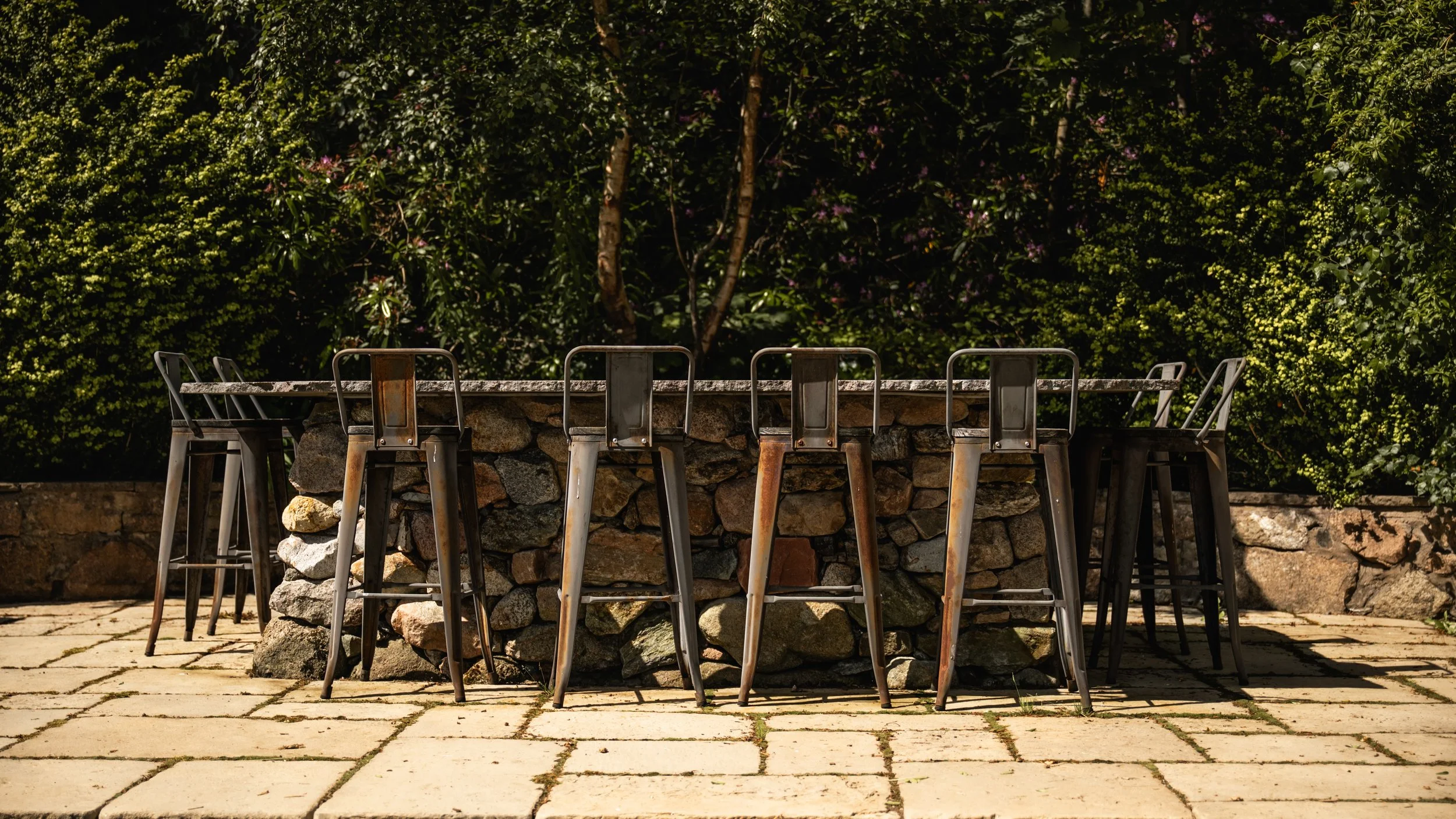 Outdoor stone table with metal chairs on a stone patio, surrounded by green bushes and trees.
