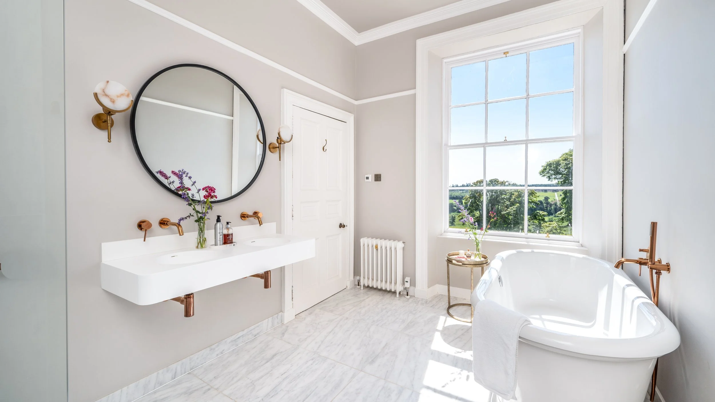 Bright bathroom with large window showing a scenic view of trees and sky, featuring a white bathtub, a double sink with copper fixtures, a mirror, and decorative flowers.