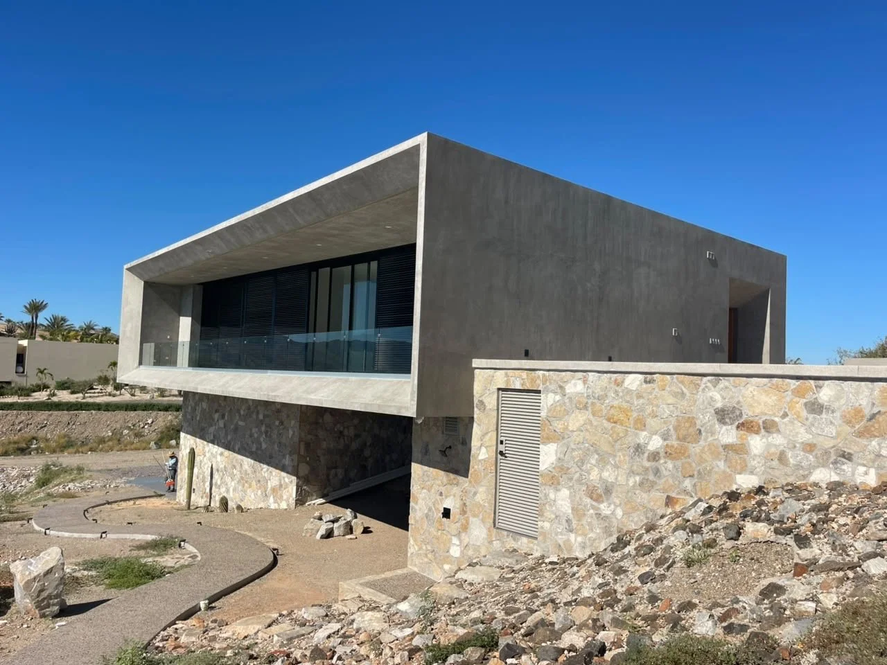 Modern house with a concrete and stone exterior, large glass balcony, located in a desert-like area with some palm trees in the background, under a clear blue sky.