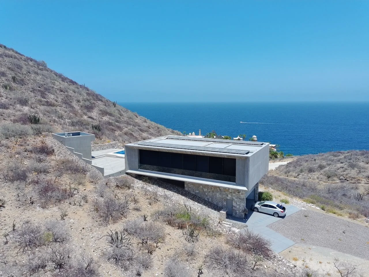 Modern house with large windows on a hillside overlooking the ocean with desert vegetation around.