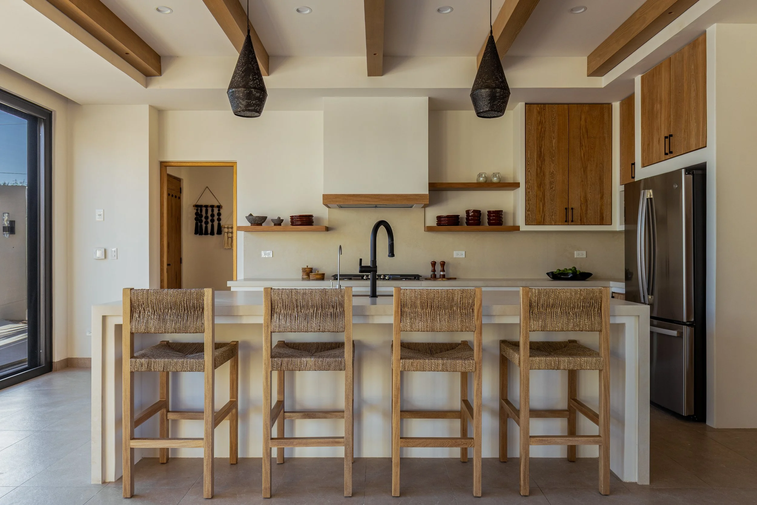 A modern kitchen with a bar-style island and four wooden chairs with woven seats, black hardware, wooden cabinets, black pendant lights, and a door leading outside.