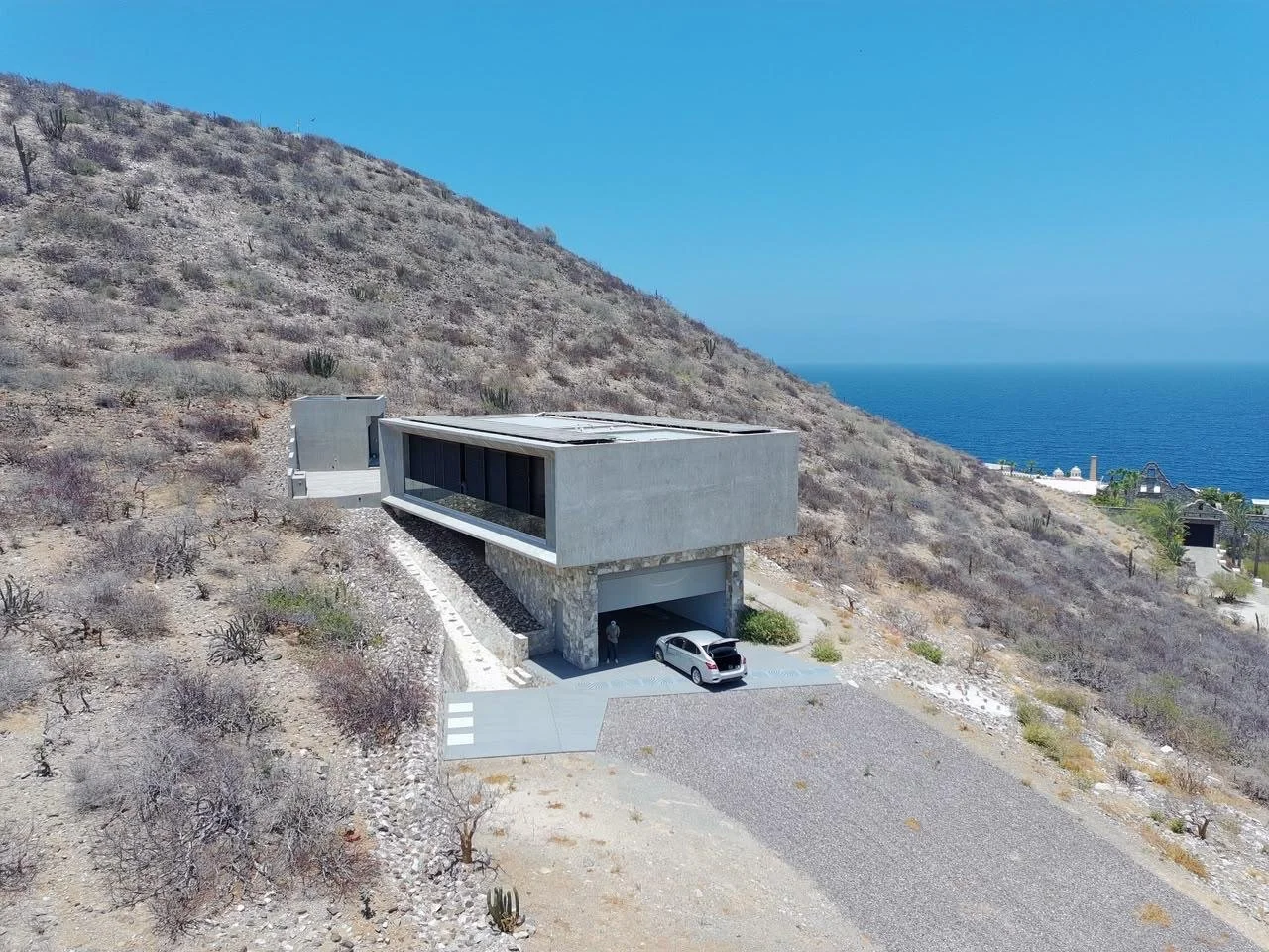 Modern house built into a hillside with a concrete exterior, overlooking the ocean in the distance. The house has a flat roof, large windows, and a garage with a car parked outside.