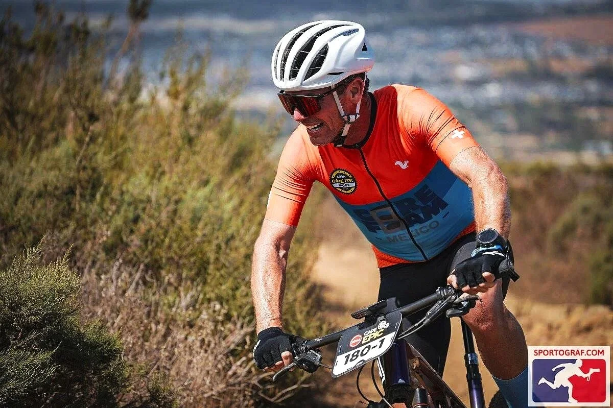 Male cyclist wearing a white helmet, red sunglasses, and an orange cycling jersey riding on a dirt trail, with a landscape in the background.