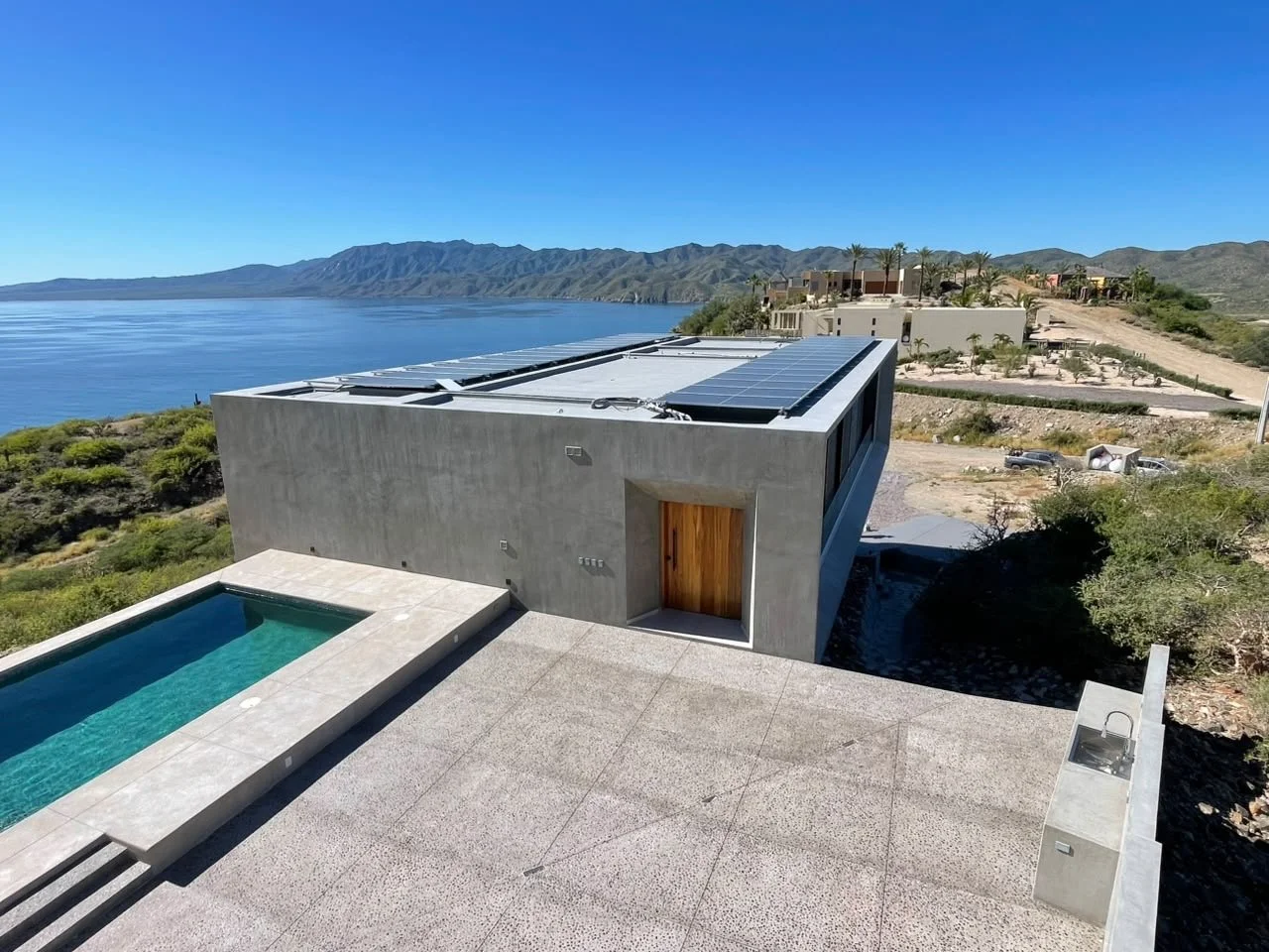 Modern concrete house with solar panels on roof, swimming pool outside, desert landscape with mountains and lake in the background.