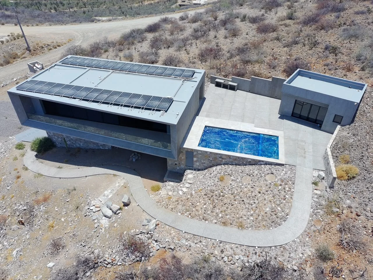 A modern house with solar panels on the roof, a swimming pool, and a curved walkway in a desert landscape.