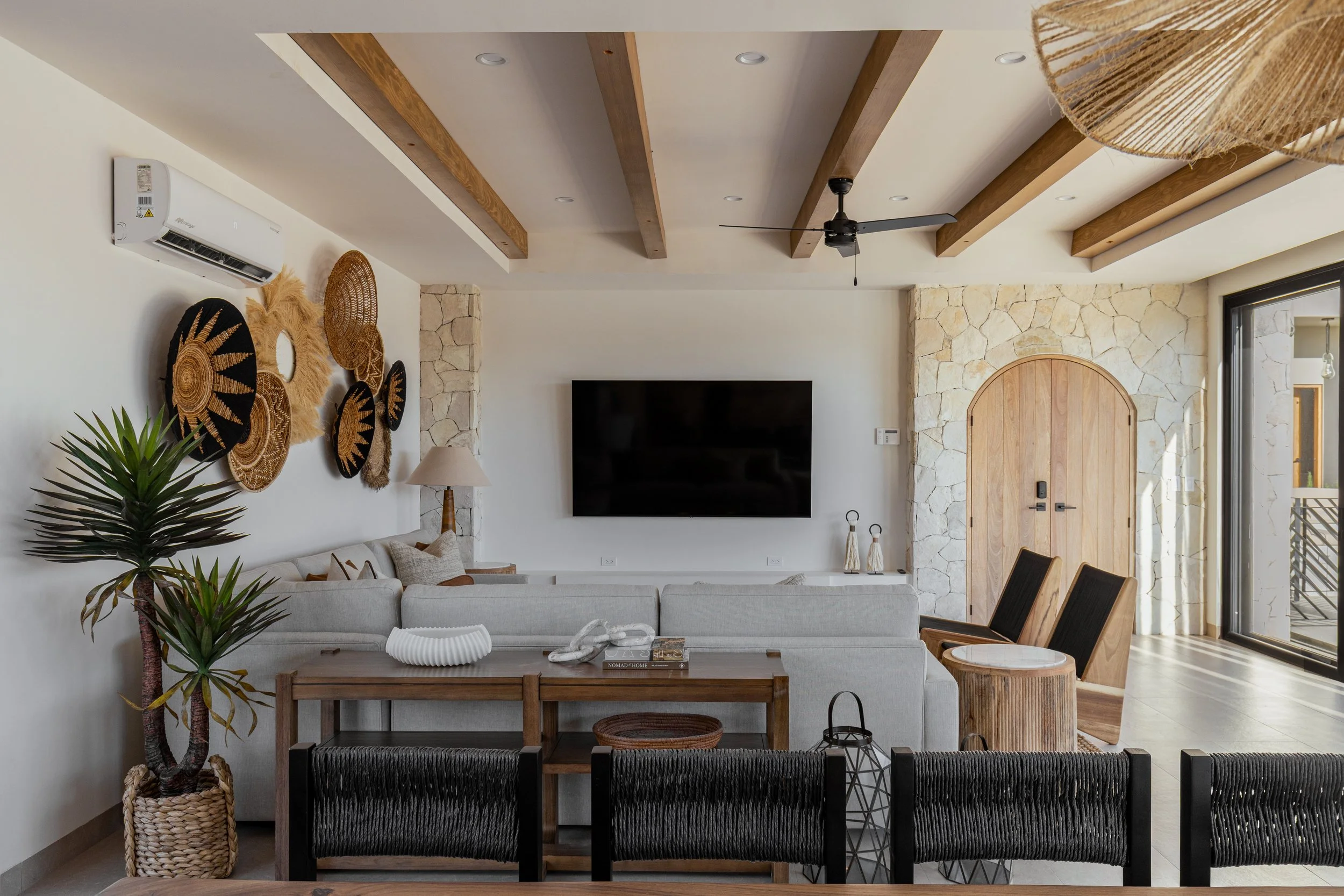 Modern living room with ceiling beams, a wall-mounted TV, a white sofa, a wooden coffee table, black chairs, a potted plant, and wall decor, with natural light coming through glass door.