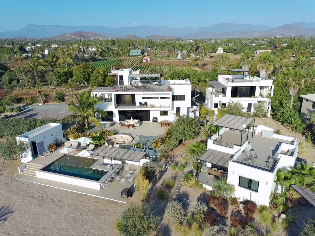 Aerial view of modern white residential buildings with rooftop terraces, swimming pool, outdoor seating, surrounded by palm trees and desert landscape.