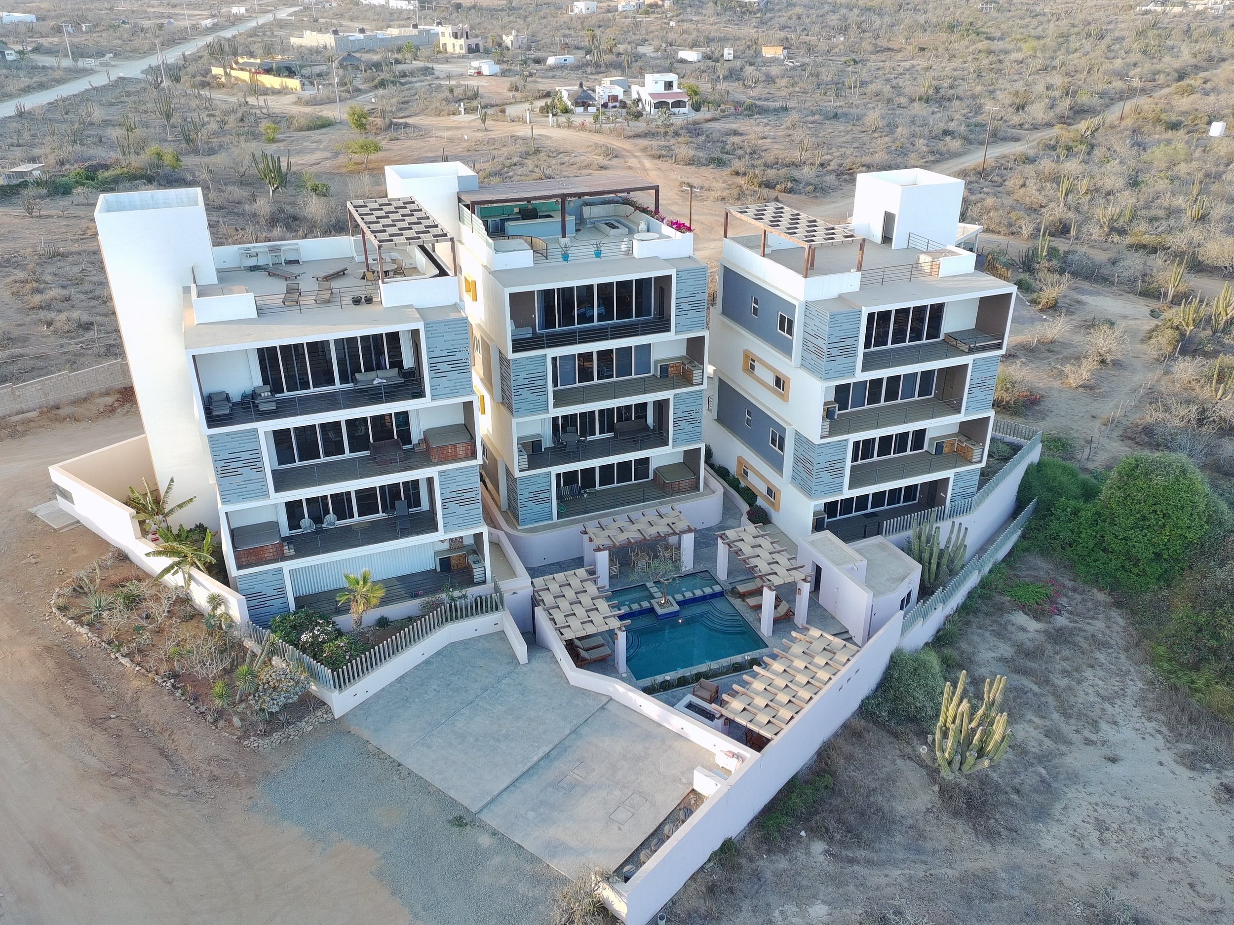 Aerial view of a modern multi-story residential building in a desert landscape with sparse vegetation and other small buildings, featuring balconies, rooftop terraces, and a swimming pool.