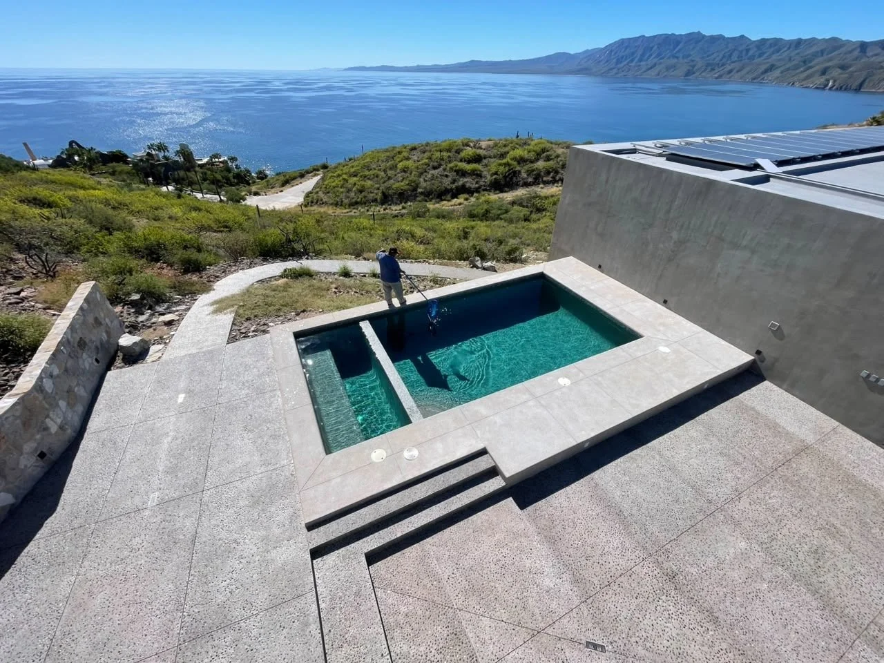 A person is cleaning a rectangular swimming pool on a terrace overlooking a scenic ocean view and distant mountains.
