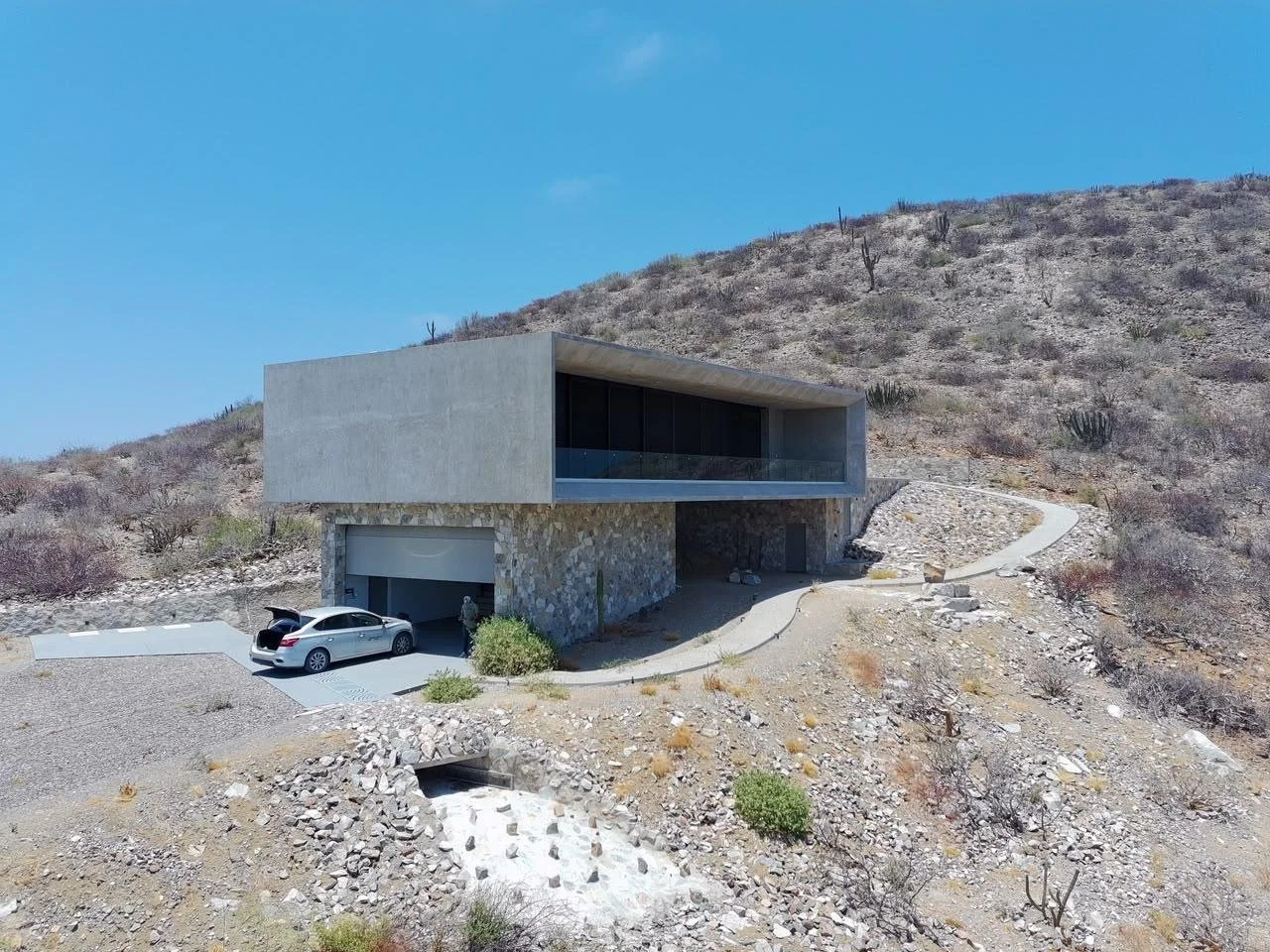 Modern house built into a hillside with a car parked in a driveway, surrounded by desert terrain and sparse vegetation.