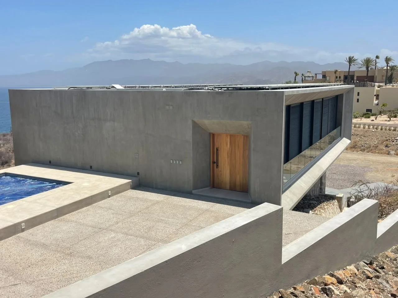 Modern concrete house with flat roof, large glass windows, wooden door, swimming pool, and a mountain view in the background.