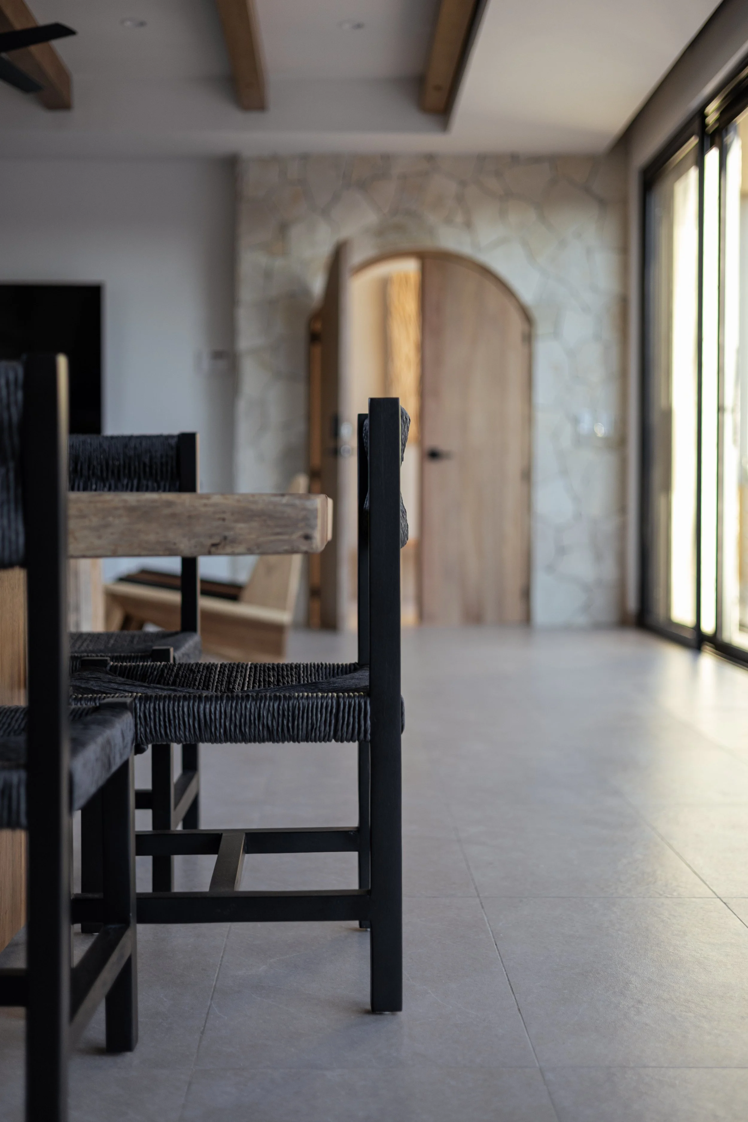 Interior view of a modern room with a stone wall, wooden ceiling beams, a large sliding glass door, and black chairs around a wooden table.