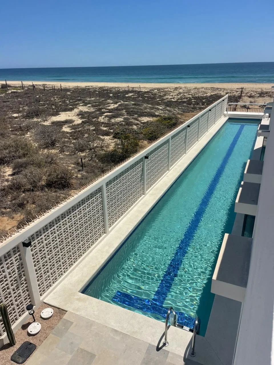 A long, narrow swimming pool on a rooftop with a white fence, overlooking a sandy beach and ocean under a clear blue sky.