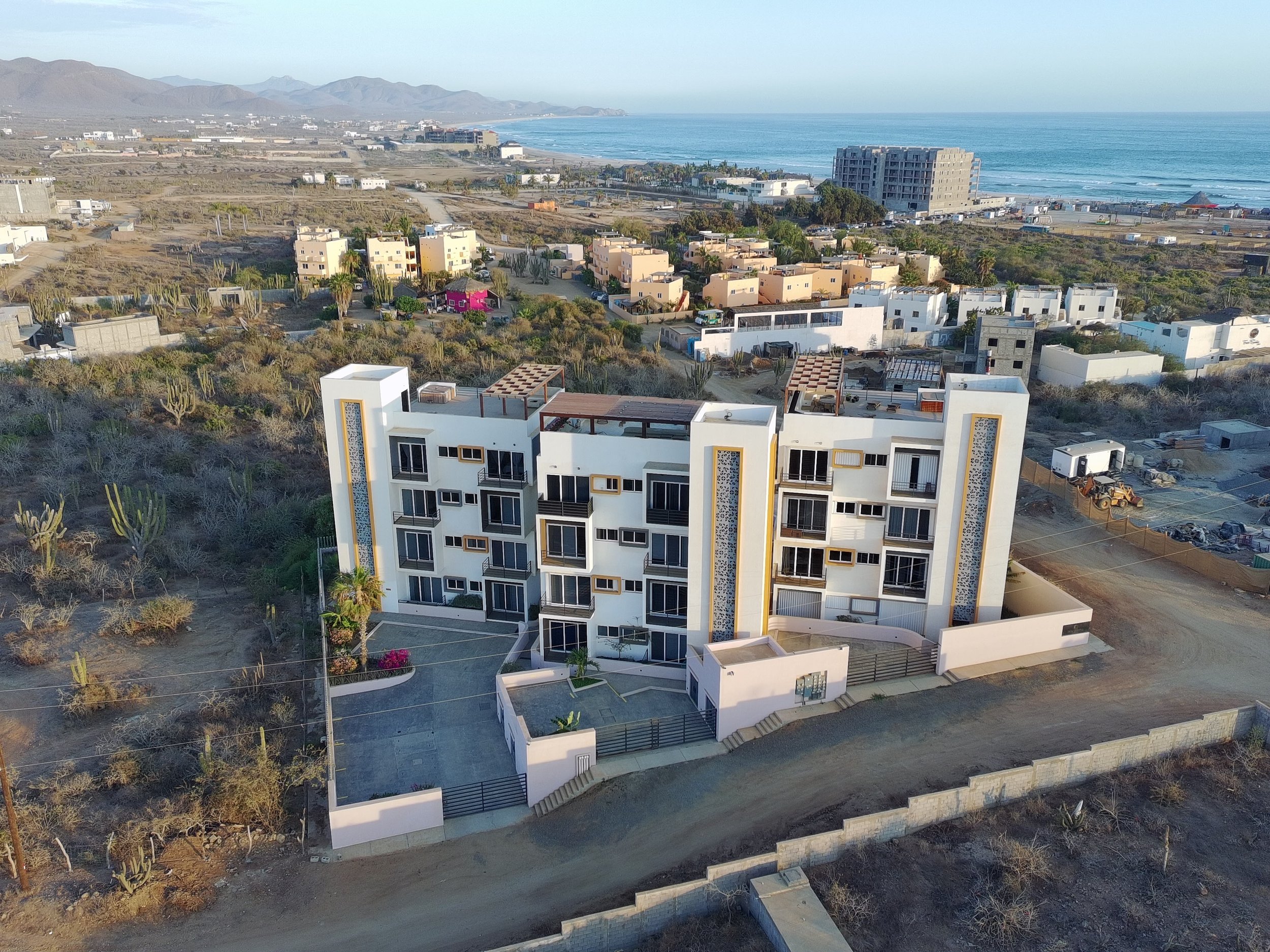 Aerial view of a coastal city with white and pastel-colored buildings, desert vegetation, and a large hotel near the ocean with sandy beaches and mountains in the background.
