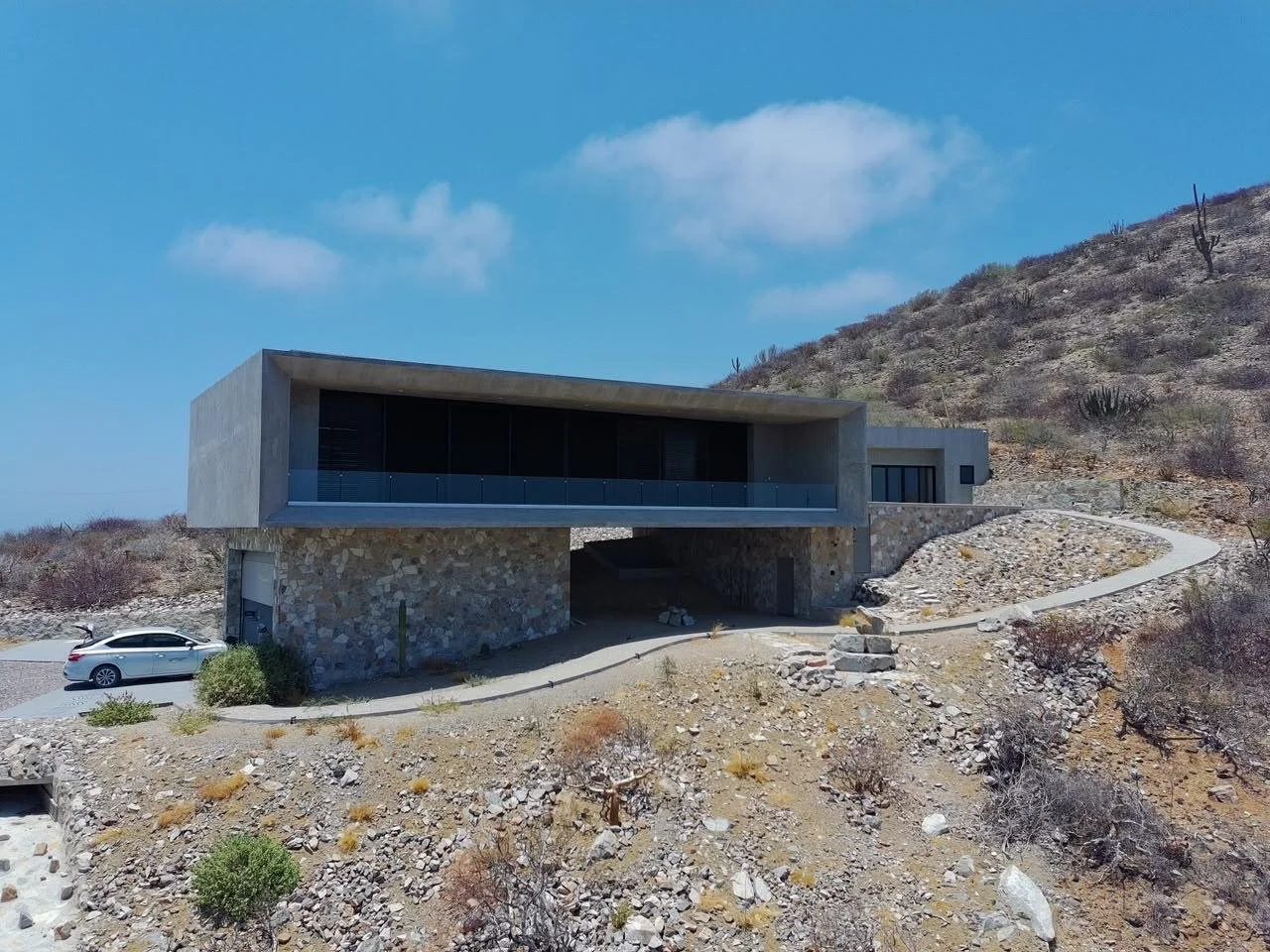 Modern house built on a hillside with stone and concrete exterior, large glass windows, and a car parked nearby in a dry, desert-like landscape with sparse vegetation and cacti under a blue sky with a few clouds.