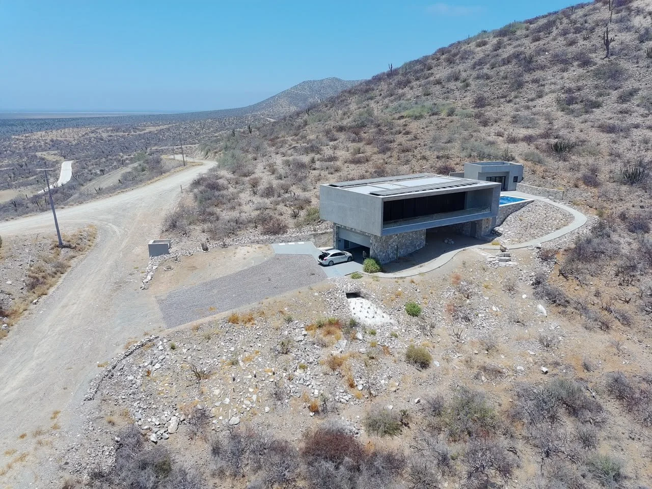 Modern house with flat roof, made of concrete and stone, situated on a desert hillside; features a small swimming pool, driveway with a white car, and a winding dirt road leading away from the house.