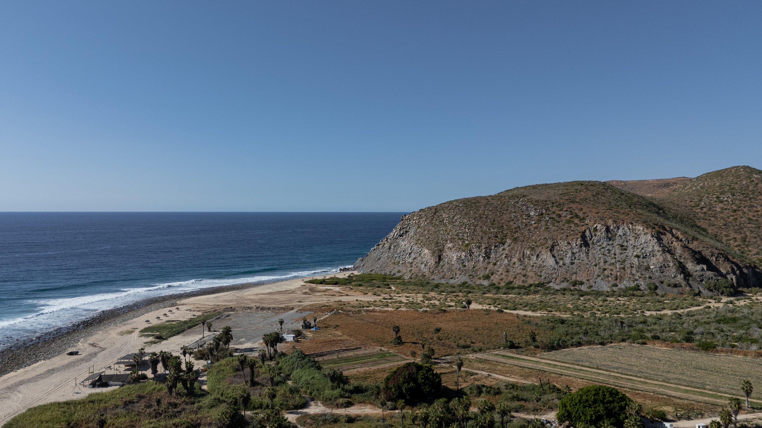 Beachside landscape with ocean, sandy beach, rocky hillside, and dry land with sparse vegetation and scattered trees under clear blue sky.