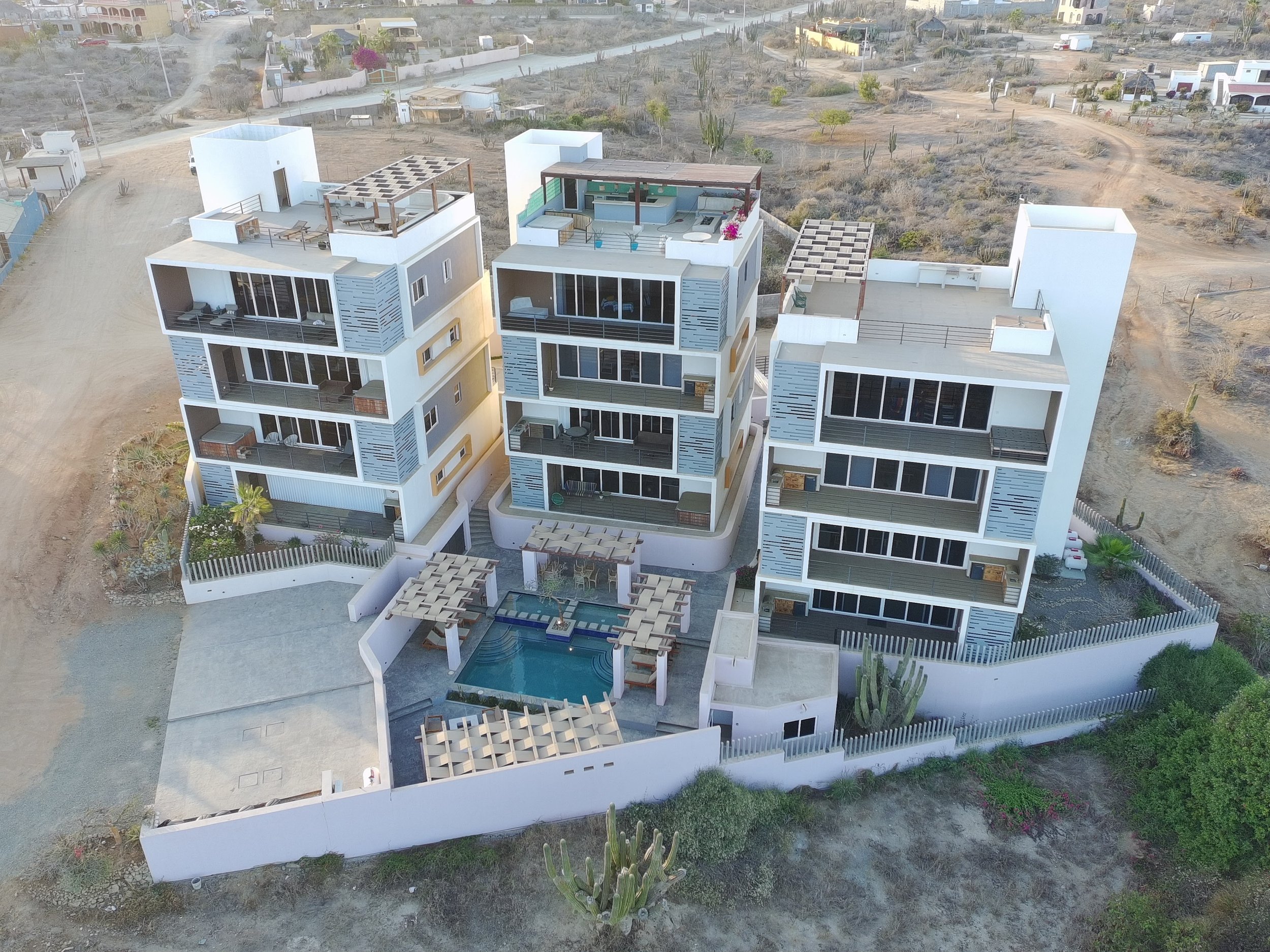 An aerial view of modern white apartment buildings with multiple balconies, some featuring outdoor furniture and rooftop gardens, in a desert landscape with sparse vegetation and cacti.